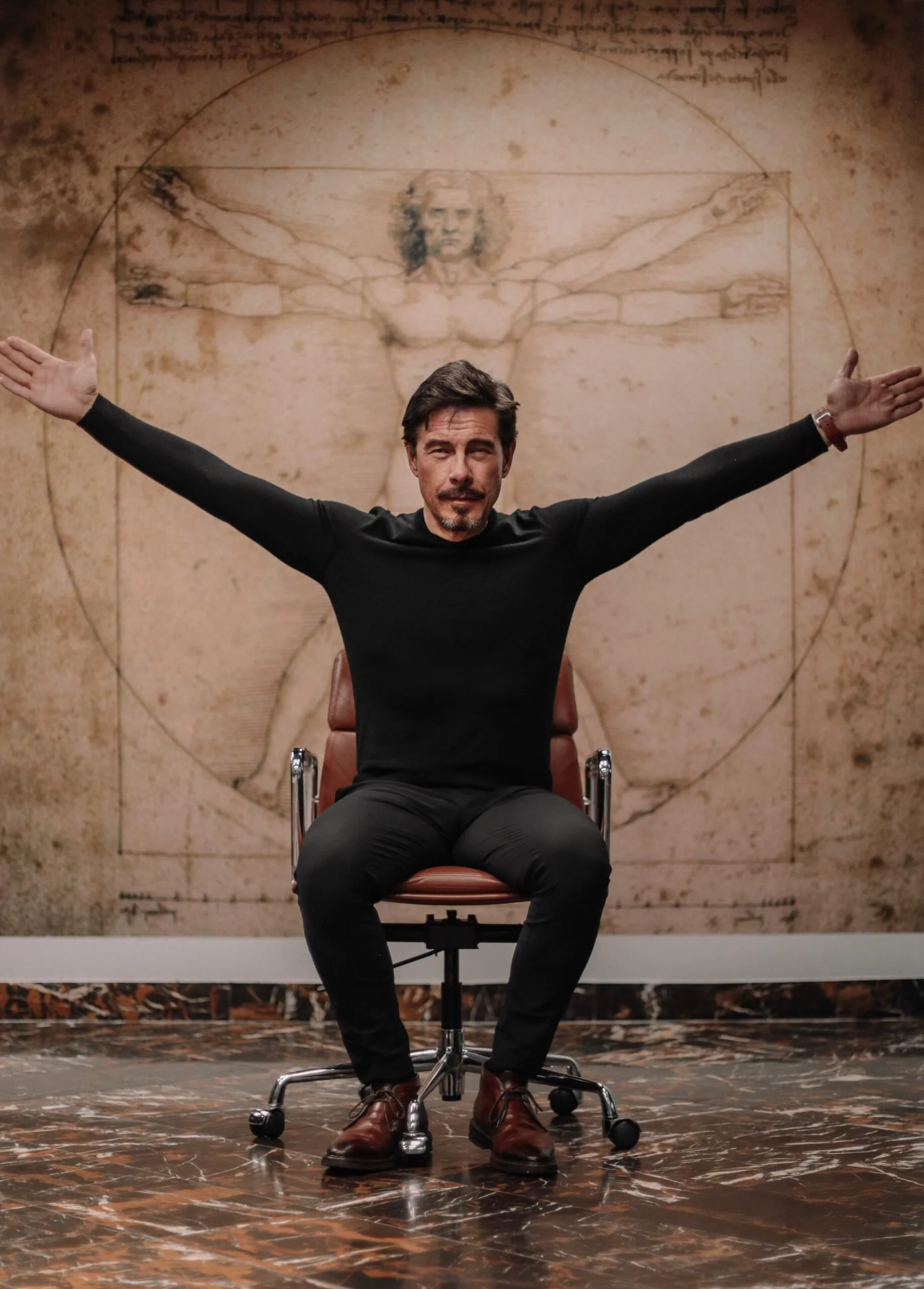 Man sitting on an office chair with arms outstretched, in front of a Leonardo da Vinci's Vitruvian Man sketch on a wall.