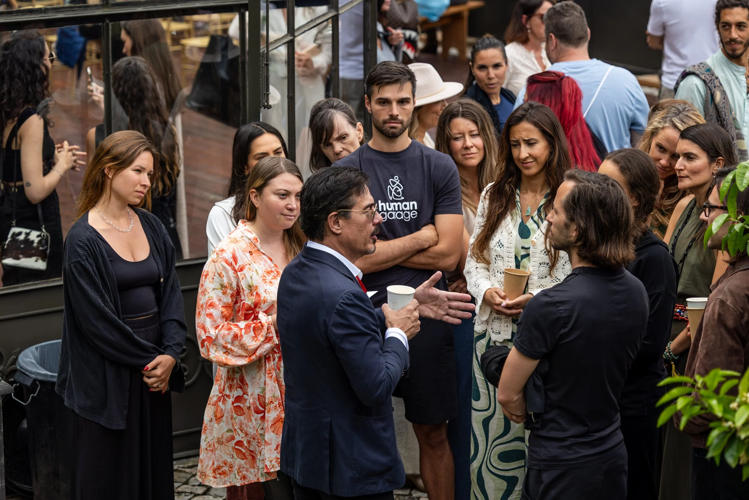 Robert E. Grant giving a speech to a group of diverse people outside, with some holding cups at the FiVth Experience in Portugal.