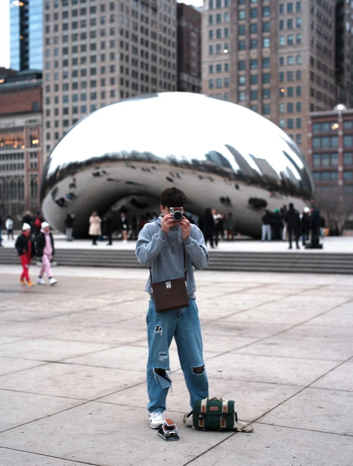 Person taking a photo in front of Cloud Gate sculpture, also known as "The Bean," in Chicago's Millennium Park, with tall buildings and people in the background.