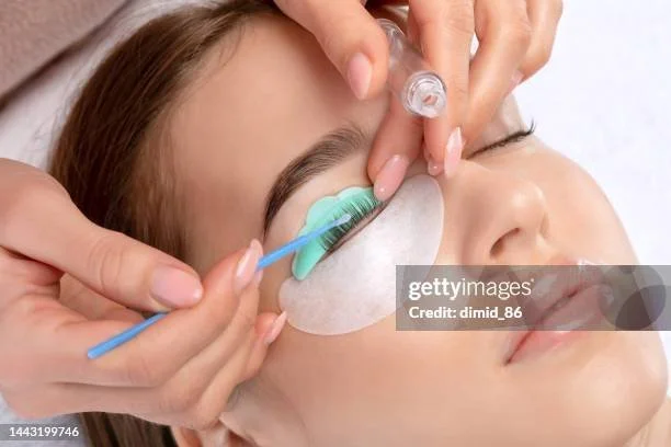 En kvinne får påsatt øyelokkforing, med en person som bruker en bomullspinne og en beholder, antagelig under en skjønnhetsbehandling.