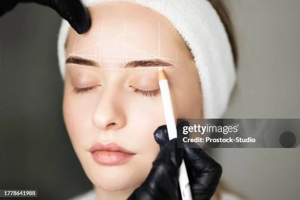 En kvinne får utført øyenbrynsskjønnhetsbehandling, med en estetiker som bruker en hvit blyant og en laser eller lysenhet.