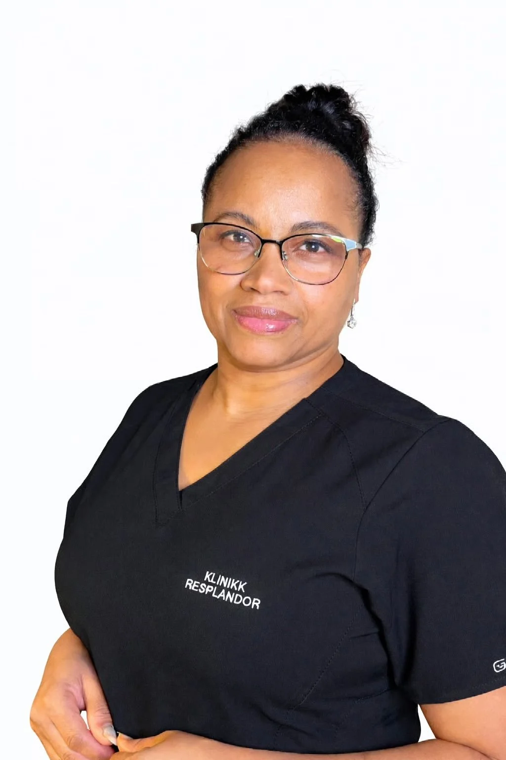 En kvinne med briller i svart medisinsk uniform med teksten "KLINIKK RESPLANDOR" over brystet, smilende og med håret oppsatt, mot en hvit bakgrunn. Klinikk. Keratinbehandling. PMU.