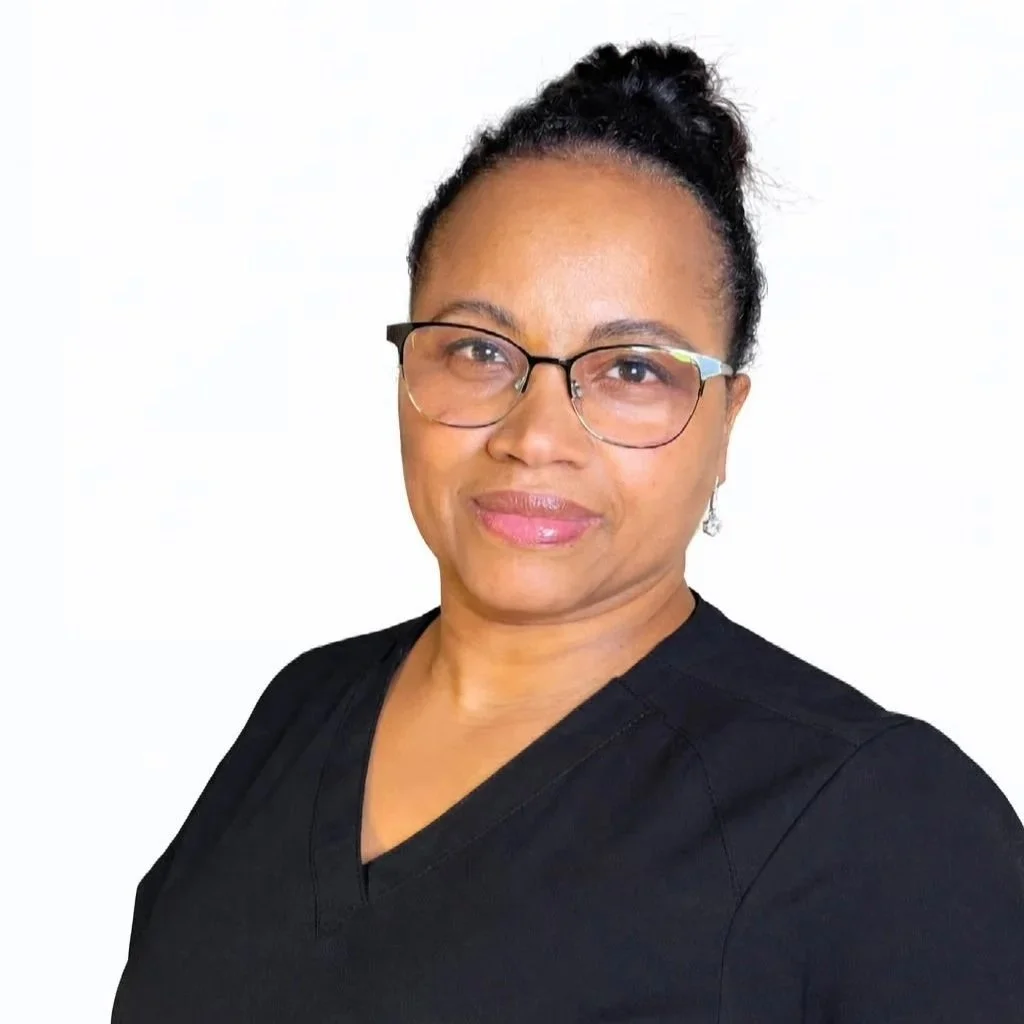 Portrett av en kvinne med briller og oppsatt hårlokk, kledd i svart uniform, mot en hvit bakgrunn. Klinikk. Keratinbehandling. PMU.