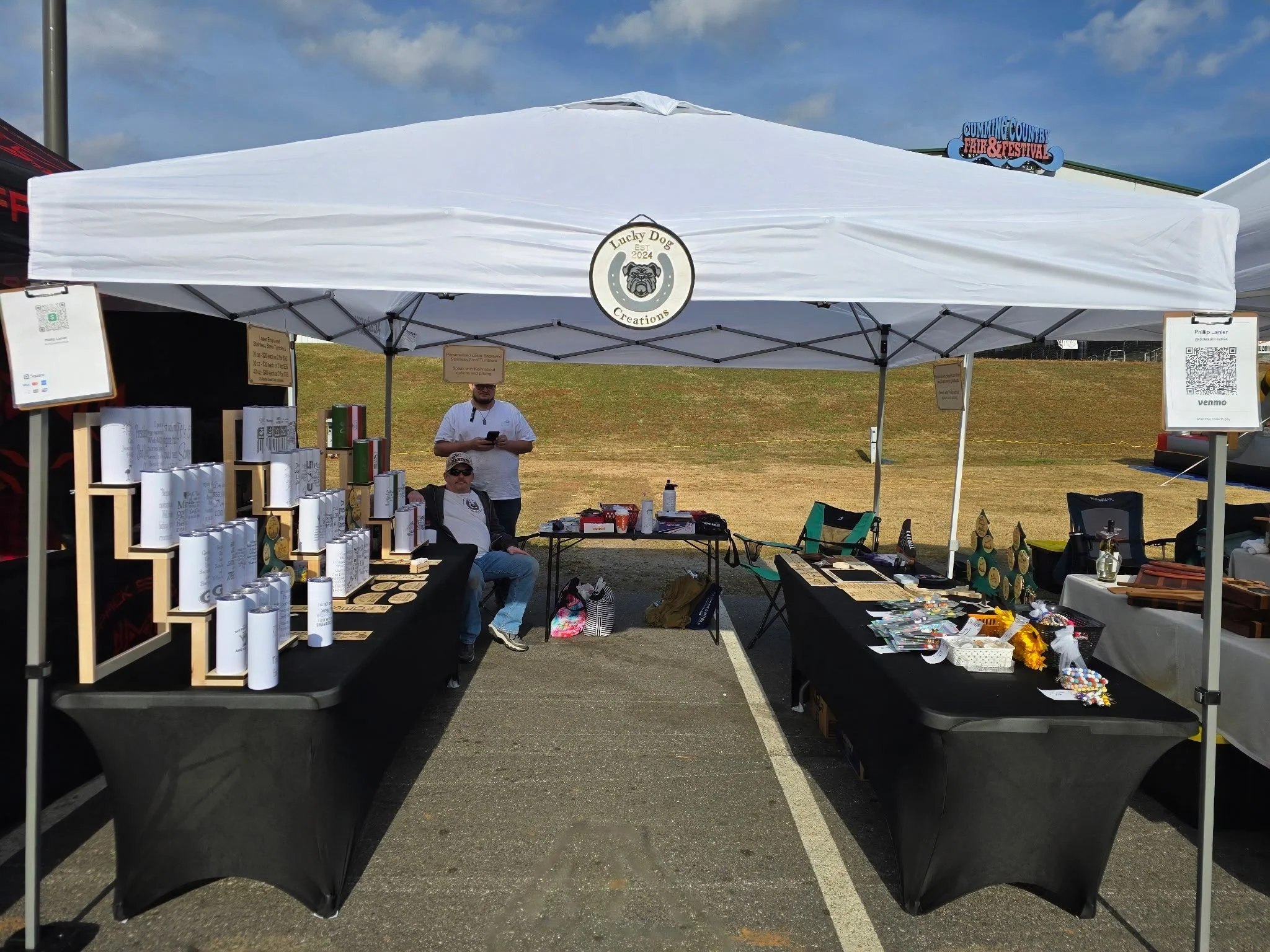 Market stall under a white tent at an outdoor event with various items on display, including crafts, and decorations, and two people sitting and standing nearby.