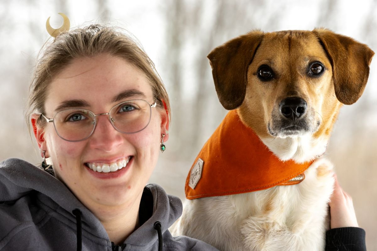A young woman with glasses and earrings smiling next to a small dog wearing an orange bandana.