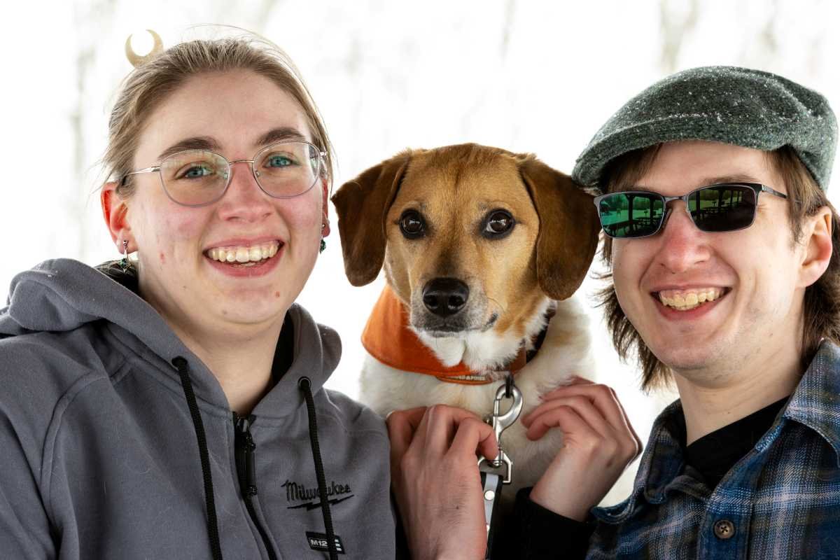Two smiling young adults, one woman and one man, hold a brown and white dog between them. The woman wears glasses and a gray hoodie; the man wears sunglasses, a gray cap, and a blue plaid shirt.