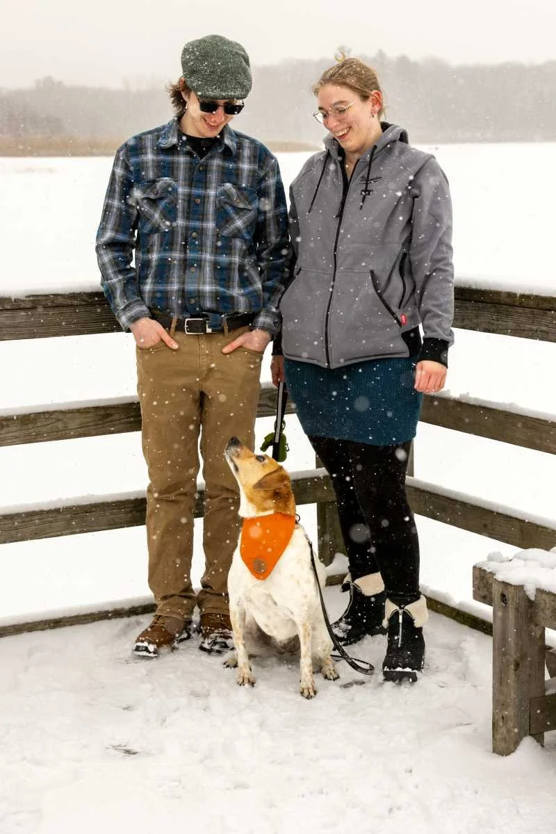 A young man and woman standing on a snowy deck with a small dog between them. The man is wearing sunglasses, a cap, and a plaid shirt, while the woman is wearing glasses, a gray hoodie, and a skirt. Snow is falling, and they are smiling at each other