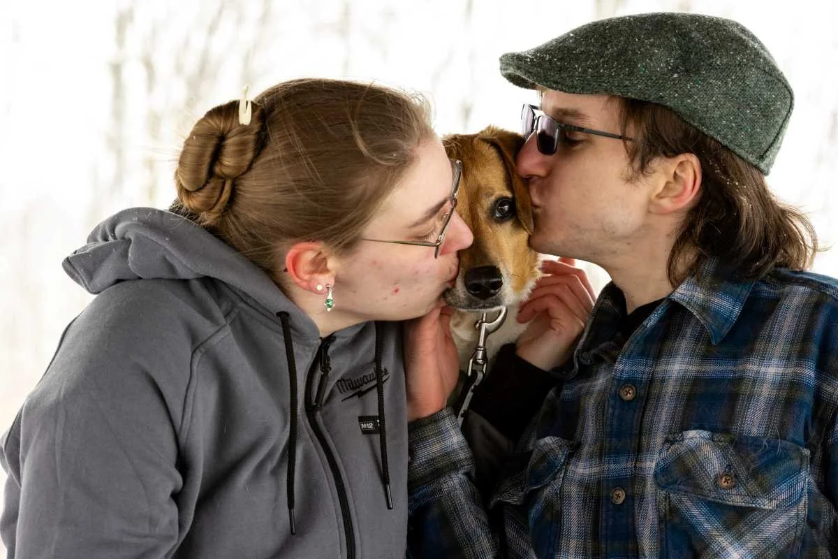 A couple kisses a dog in the middle of their faces, with the woman wearing glasses and a gray hoodie, and the man wearing glasses, a gray cap, and a plaid shirt, celebrating Valentine's Day in a bright, outdoor setting.