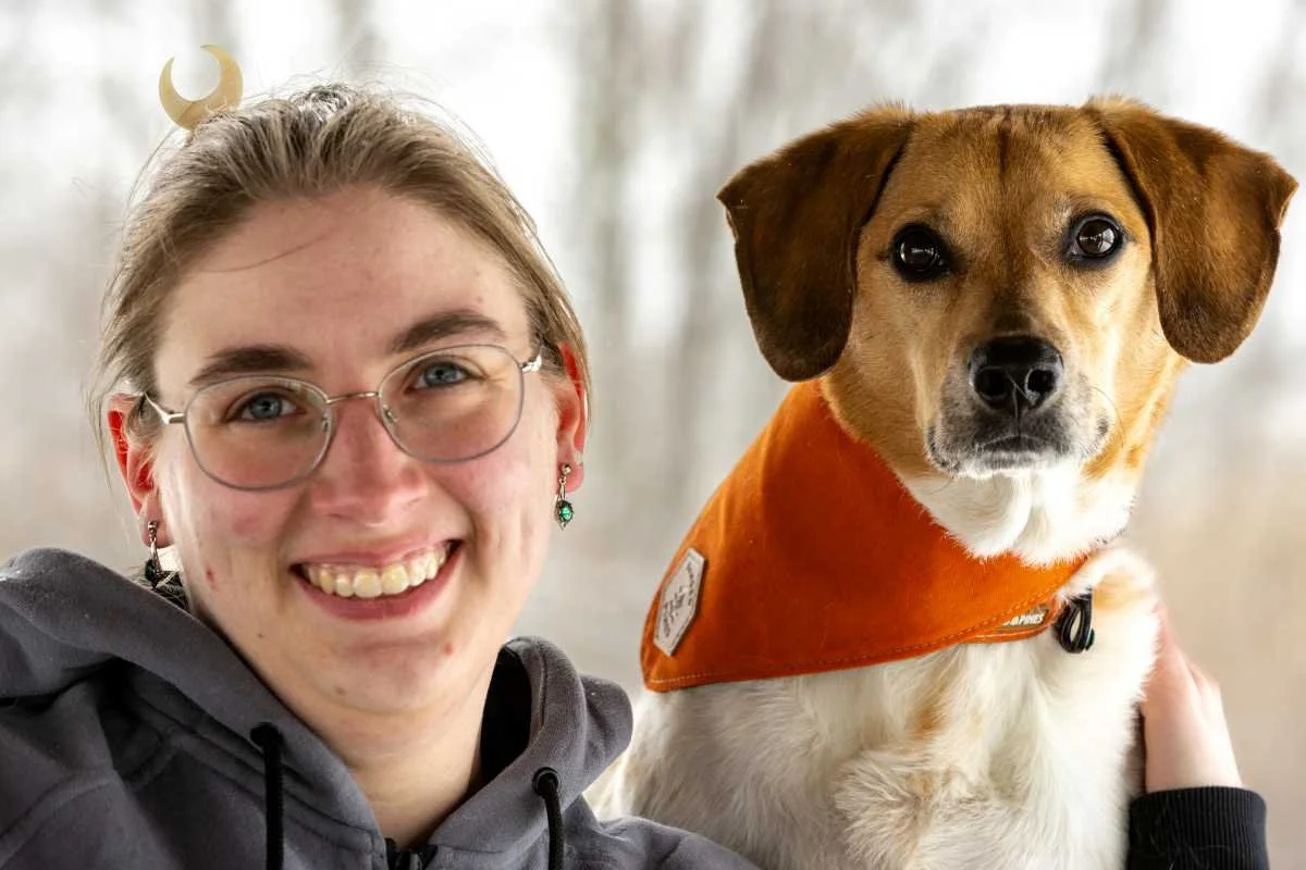 A young woman with glasses and earrings, smiling, sitting next to a dog with a brown and white coat, black eyes, and wearing an orange bandana.