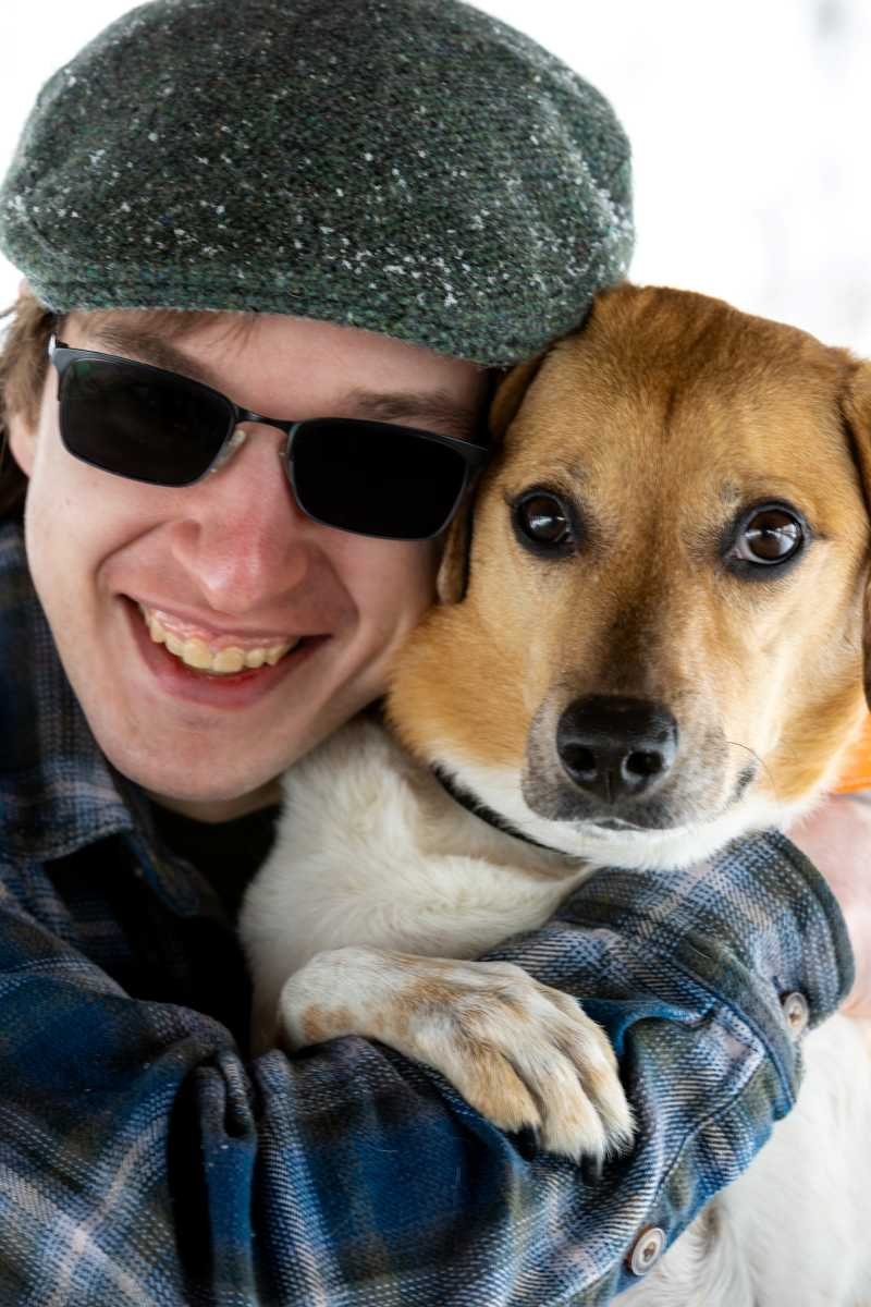 A man wearing sunglasses, a gray hat, and a plaid shirt hugging a brown and white dog with expressive eyes.