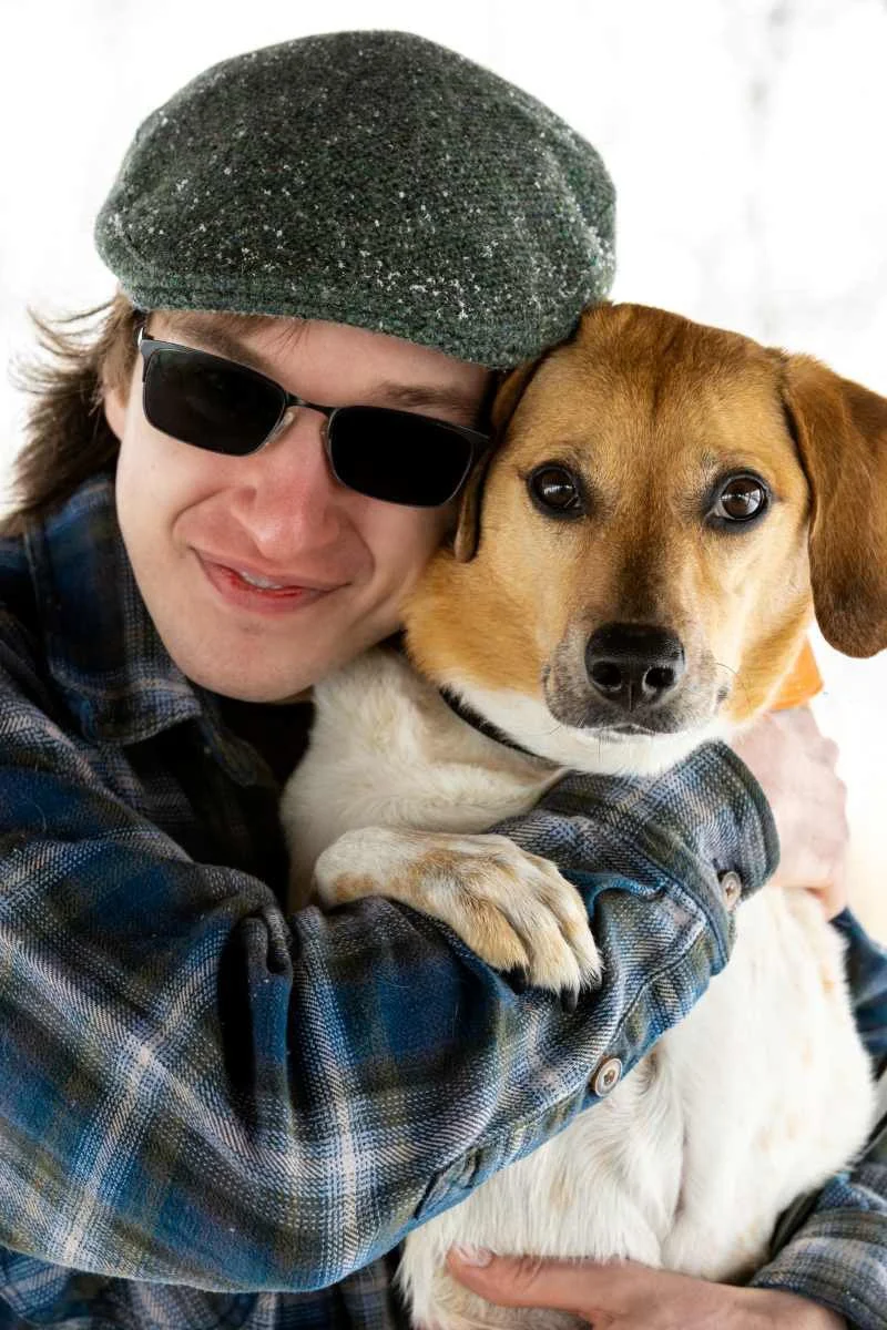 A person wearing a green beret and sunglasses hugging a brown and white dog.