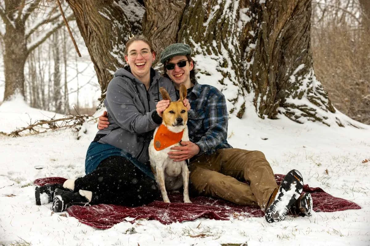 A happy couple with their dog sitting on a red blanket in a snowy outdoor setting near a large tree.