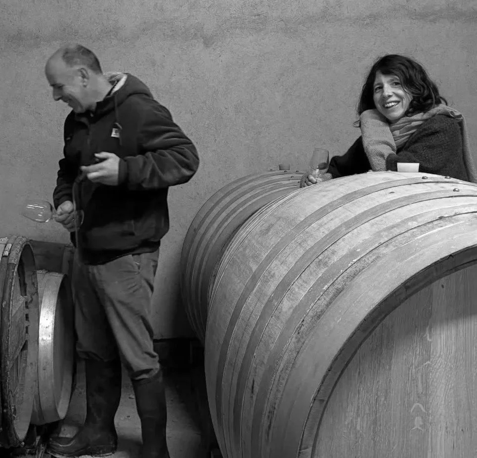 A woman sitting on a large wine barrel, smiling and holding a glass of wine. A man standing nearby, holding a glass of wine, in a winery setting.