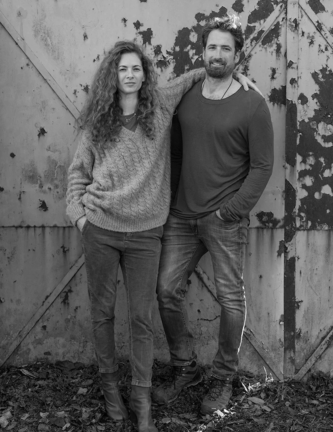 A black-and-white photo of a man and woman standing close together, with the woman resting her arm on the man's shoulder. They are in front of a weathered, peeling metal wall, dressed casually.