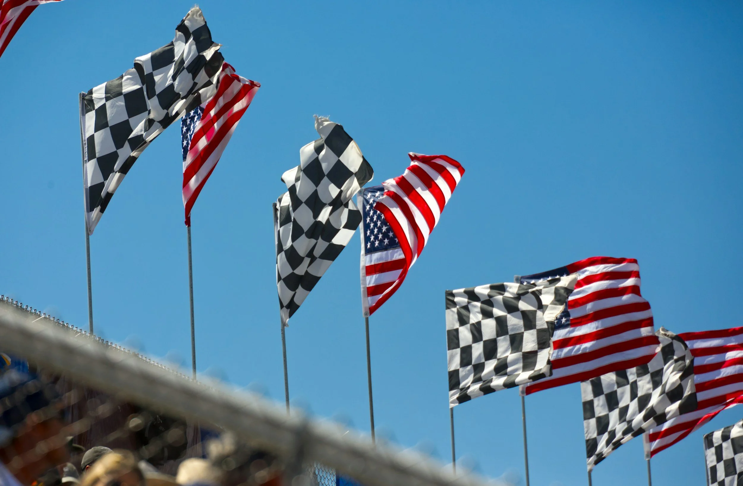 American flags and checkered flags waving against a bright blue sky over a nascar race track.
