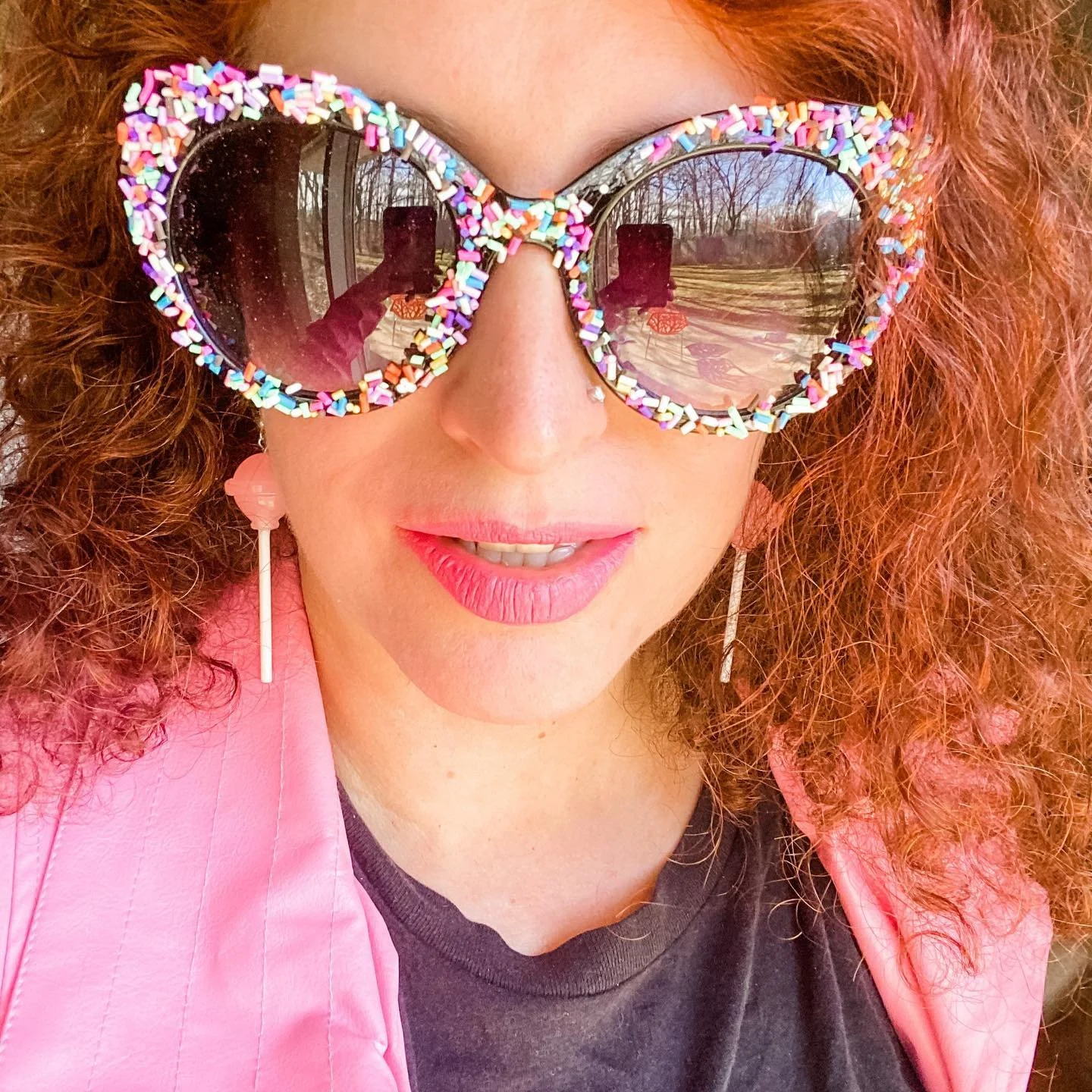 A woman with curly red hair wearing large sunglasses adorned with colorful sprinkles, pink lipstick, a pink jacket, and a black shirt, standing outdoors on a sunny day.