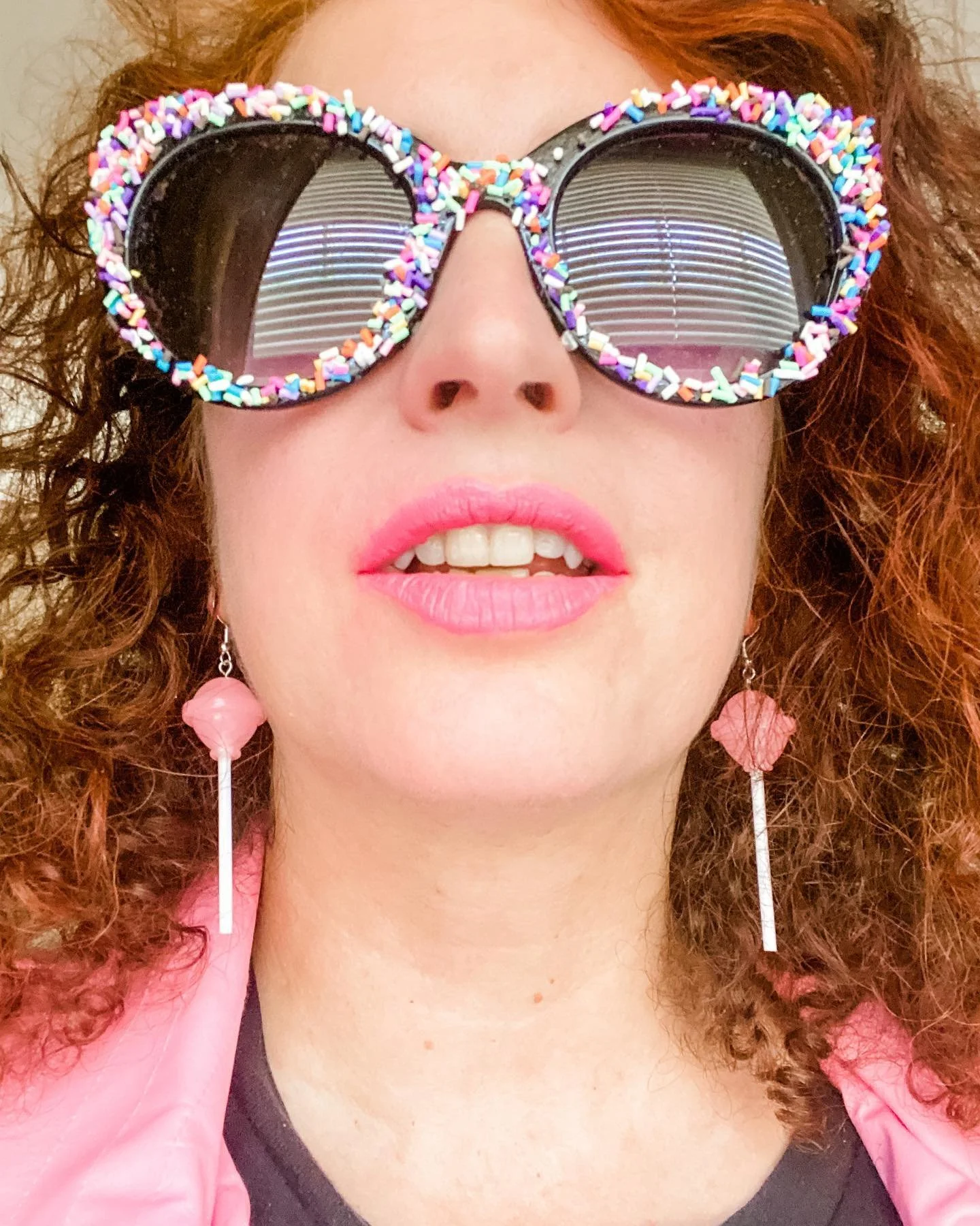 Close-up of a woman wearing oversized sunglasses decorated with colorful sprinkles, pink lipstick, pink earrings, and a pink jacket.