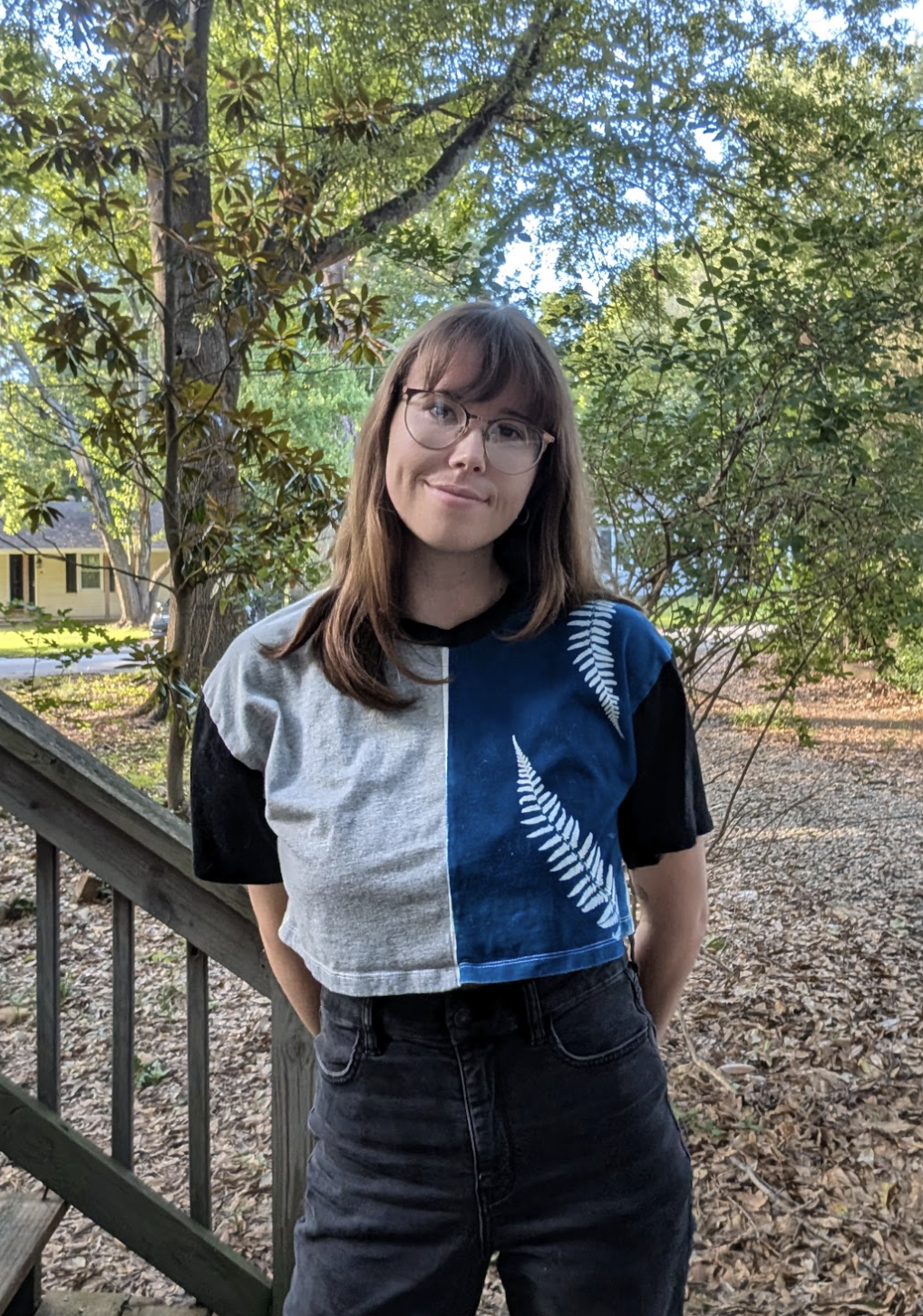 A woman with shoulder-length brown hair and glasses standing outdoors in a wooded area during daytime, wearing a gradient-colored crop top with a fern design and black high-waisted jeans, smiling softly.