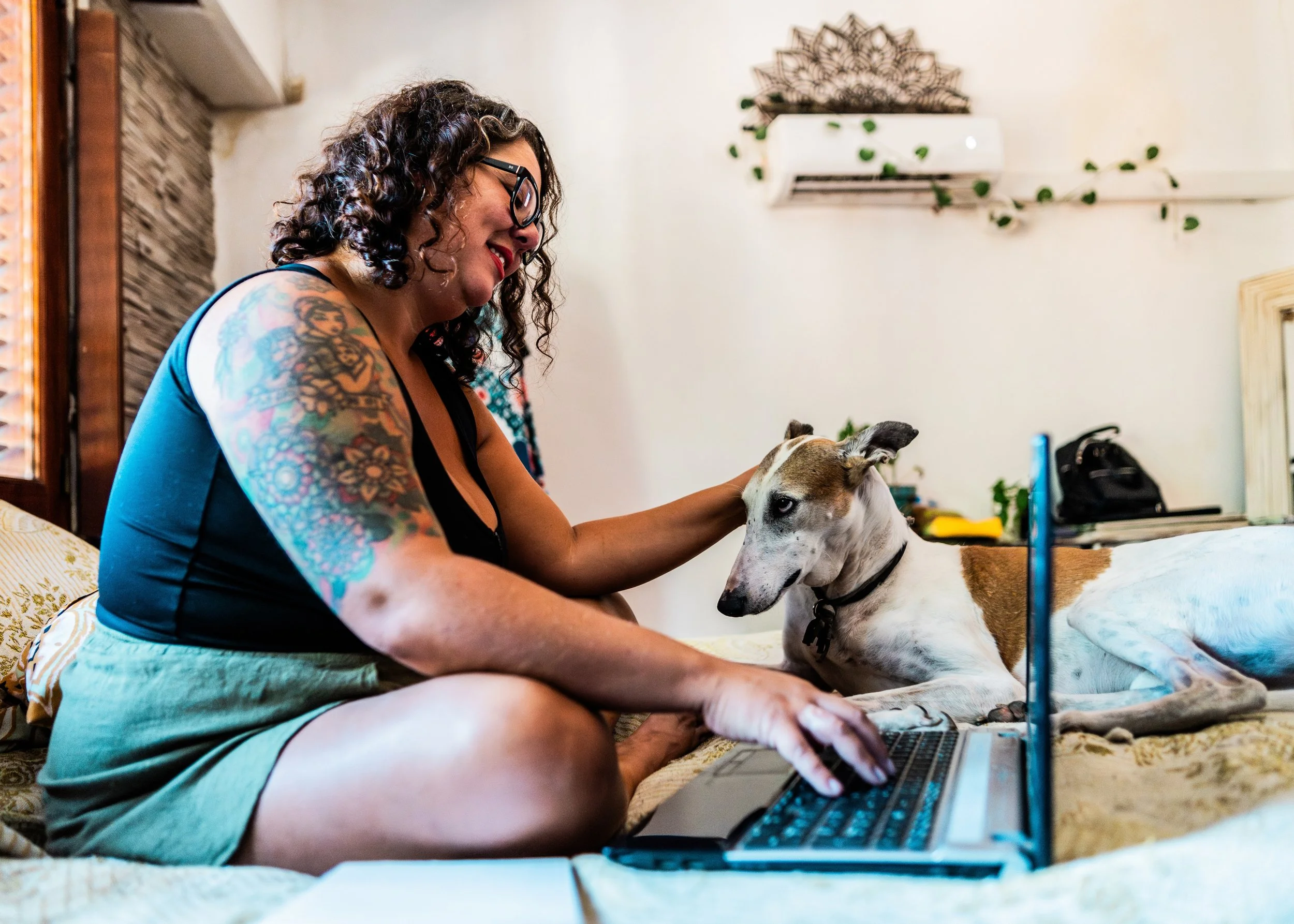 Person with curly hair and tattoos sitting on a bed using a laptop, petting a dog. The room is decorated with plants and a wall sculpture.