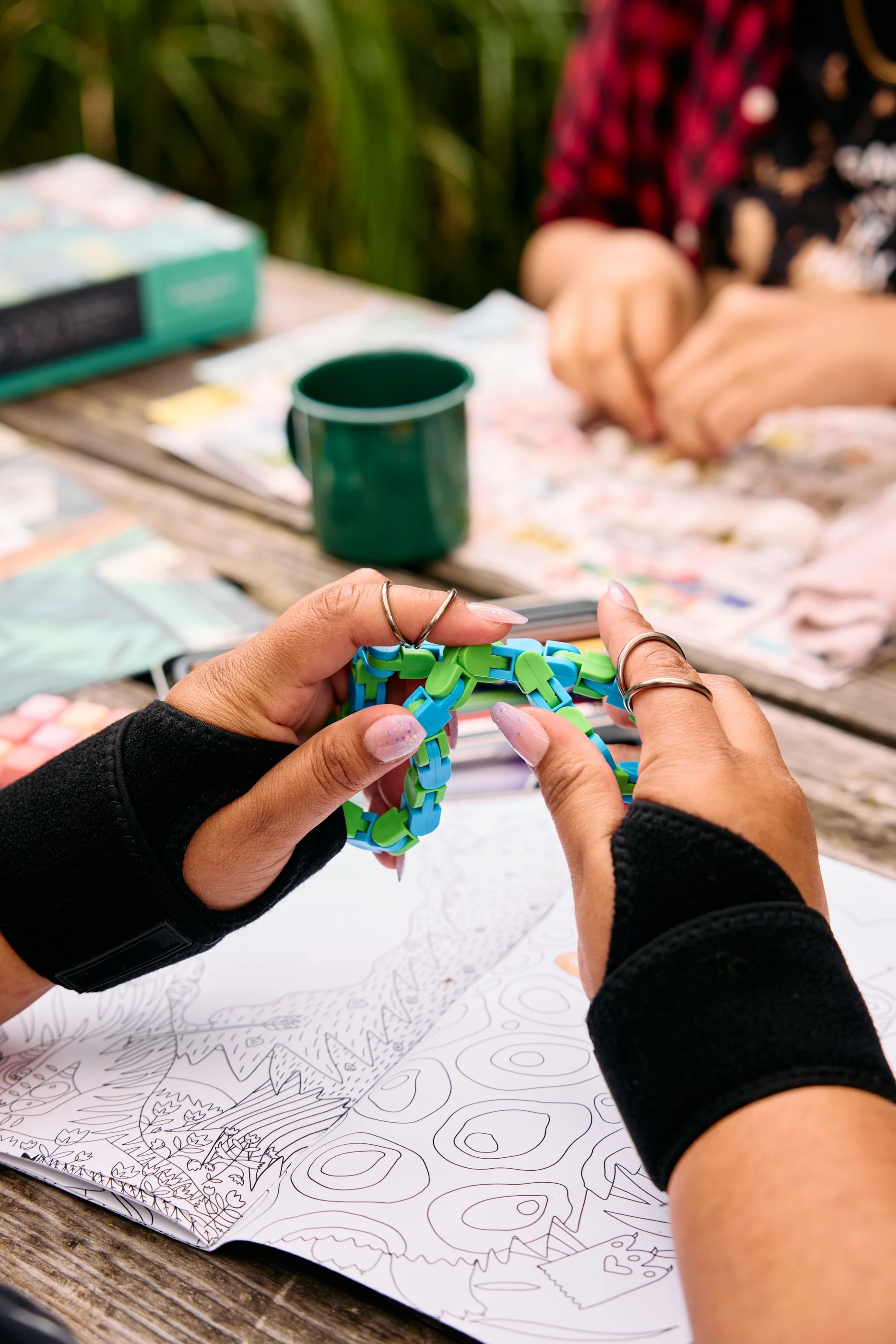 Person twisting a colorful chain link bracelet with a coloring book and art supplies on a wooden table outdoors.