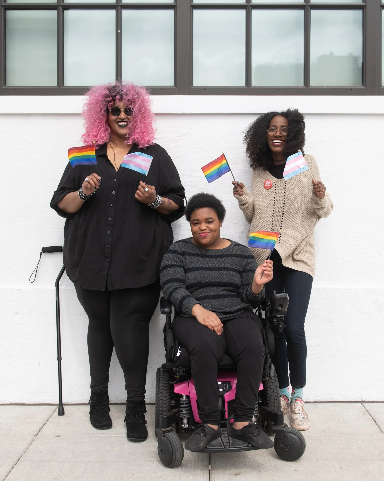 Three people celebrating LGBTQ+ pride, holding rainbow and trans pride flags, standing against a white wall outdoors, one person in a wheelchair, the other two standing, smiling and wearing casual clothing and accessories.