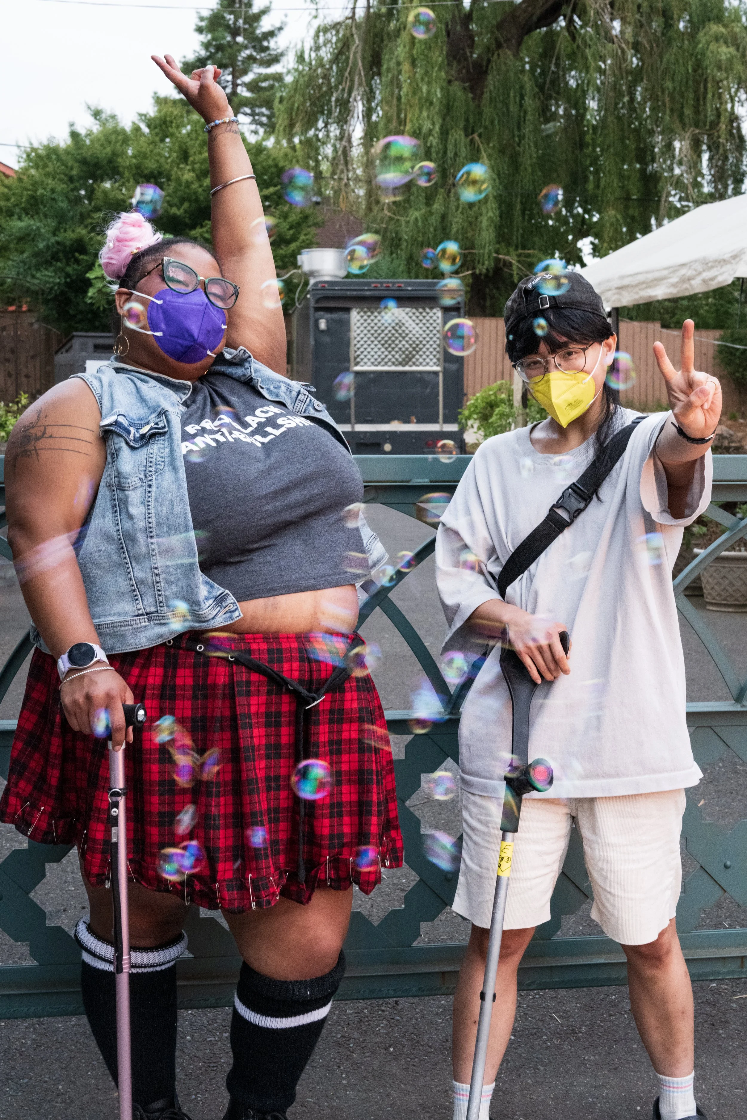 Two people, both wearing face masks, standing outdoors amidst floating bubbles. The person on the left is a plus-sized individual with pink hair tied in a bun, wearing glasses, a purple mask, a denim vest, a black t-shirt, a plaid red skirt, and knee-high socks. The person on the right is a slimmer individual with short black hair, glasses, a yellow mask, a loose white t-shirt, beige shorts, and a black crossbody bag, flashing peace signs with their hands.