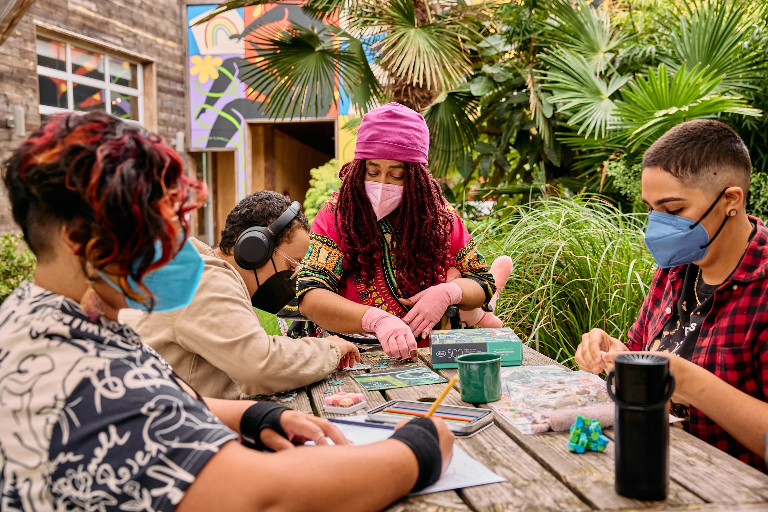 A group of people wearing face masks and gloves engaging in a tabletop activity outdoors, surrounded by lush greenery and colorful artwork.