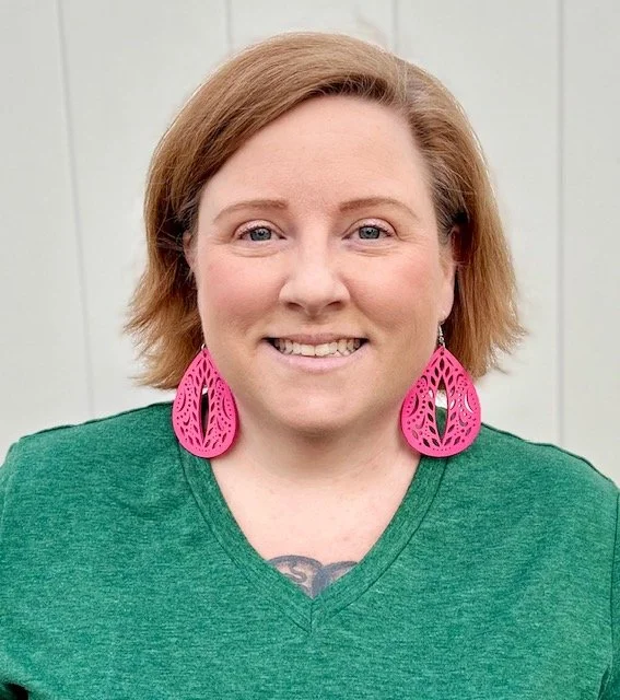 A woman with short brown hair smiles at the camera. She is wearing a green v-neck shirt and large, pink, intricately designed earrings. A small tattoo is partially visible at the top of her chest. The background is neutral, off white.
