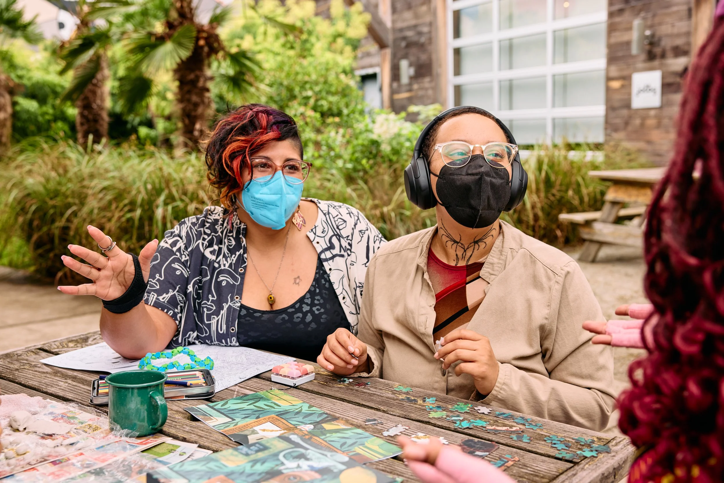 Group of people wearing face masks, engaging in a conversation outdoors at a wooden table with puzzle pieces, coloring supplies, and artwork, surrounded by green plants and trees.