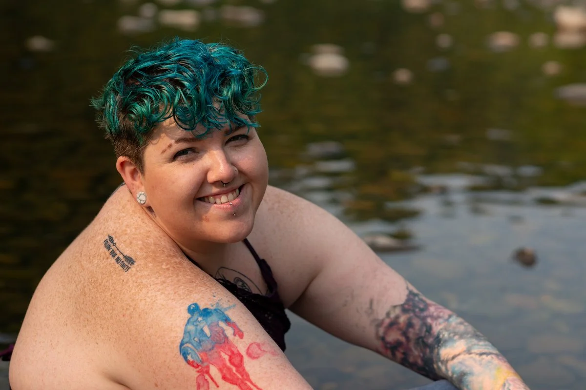 A smiling person with teal and curly hair in a water setting, showing tattoos on her shoulders and arms.