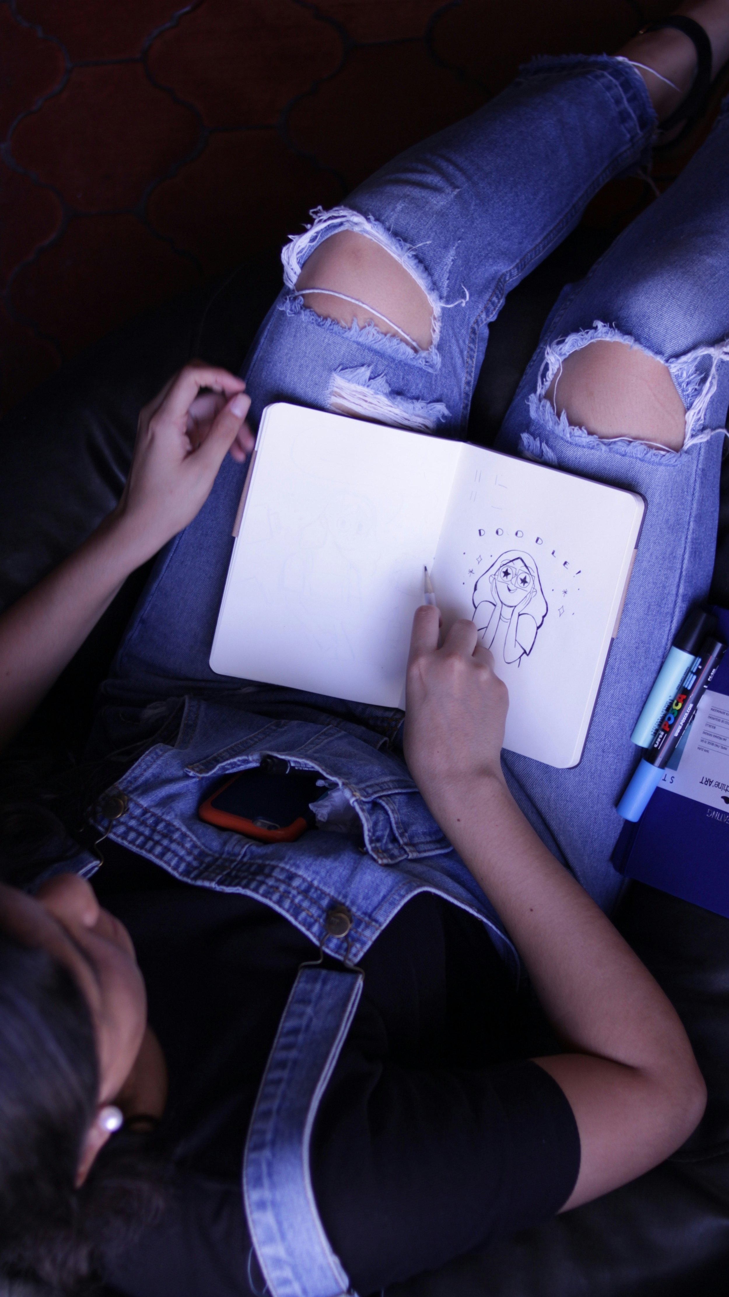 Overhead view of a person sitting on a black leather couch with ripped jeans, sketching in a notebook. The sketch is of a girl with glasses, and the word 'Doodle!' written above her. Items around include a blue folder, two markers, and a cellphone in the person's pocket.