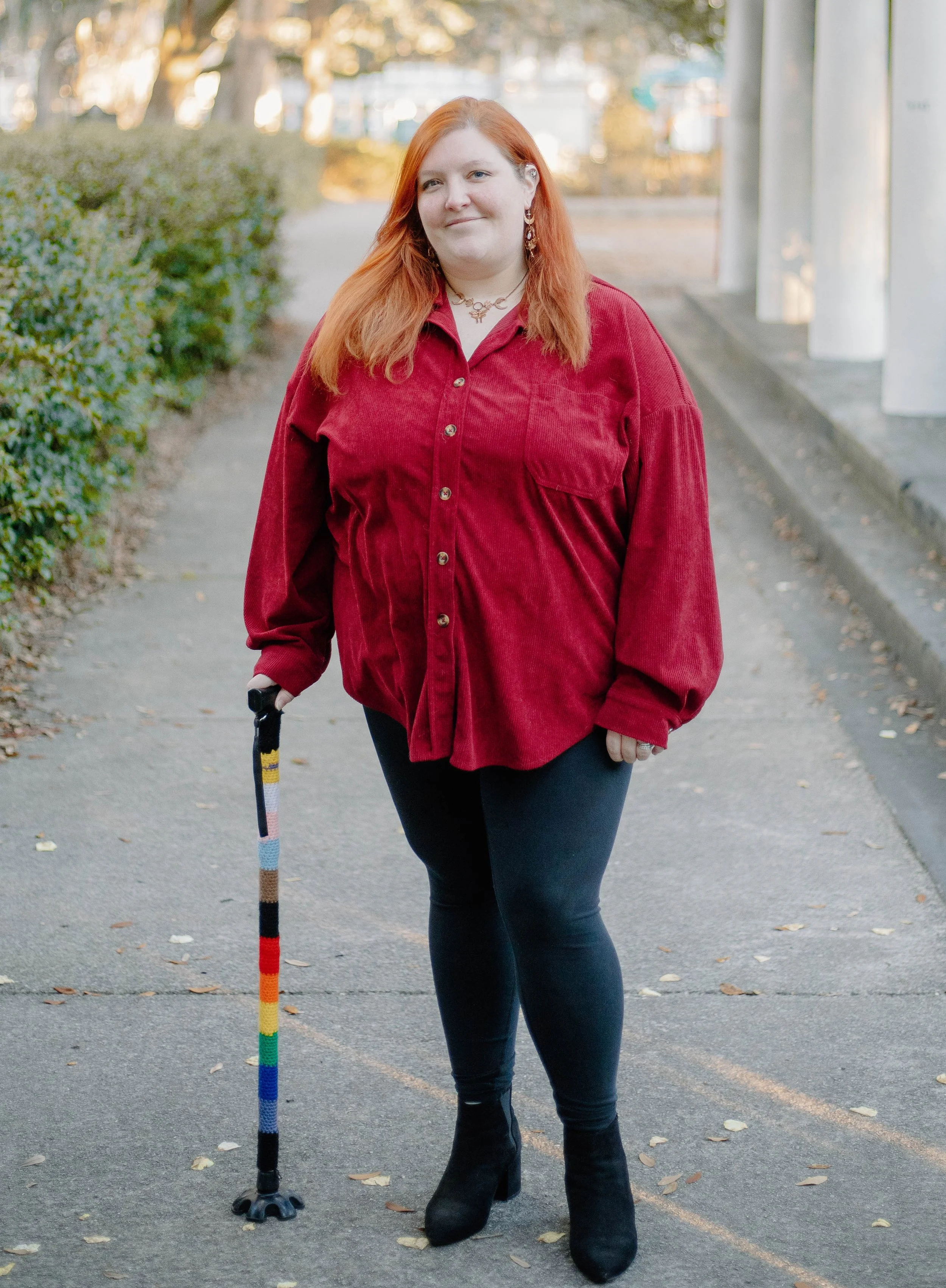 A woman with red hair and a warm smile stands on a scenic pathway lined with lush greenery and white columns. She is wearing a deep red button-up shirt, black leggings, and black boots while holding a cane wrapped in a crochet progress pride flag.