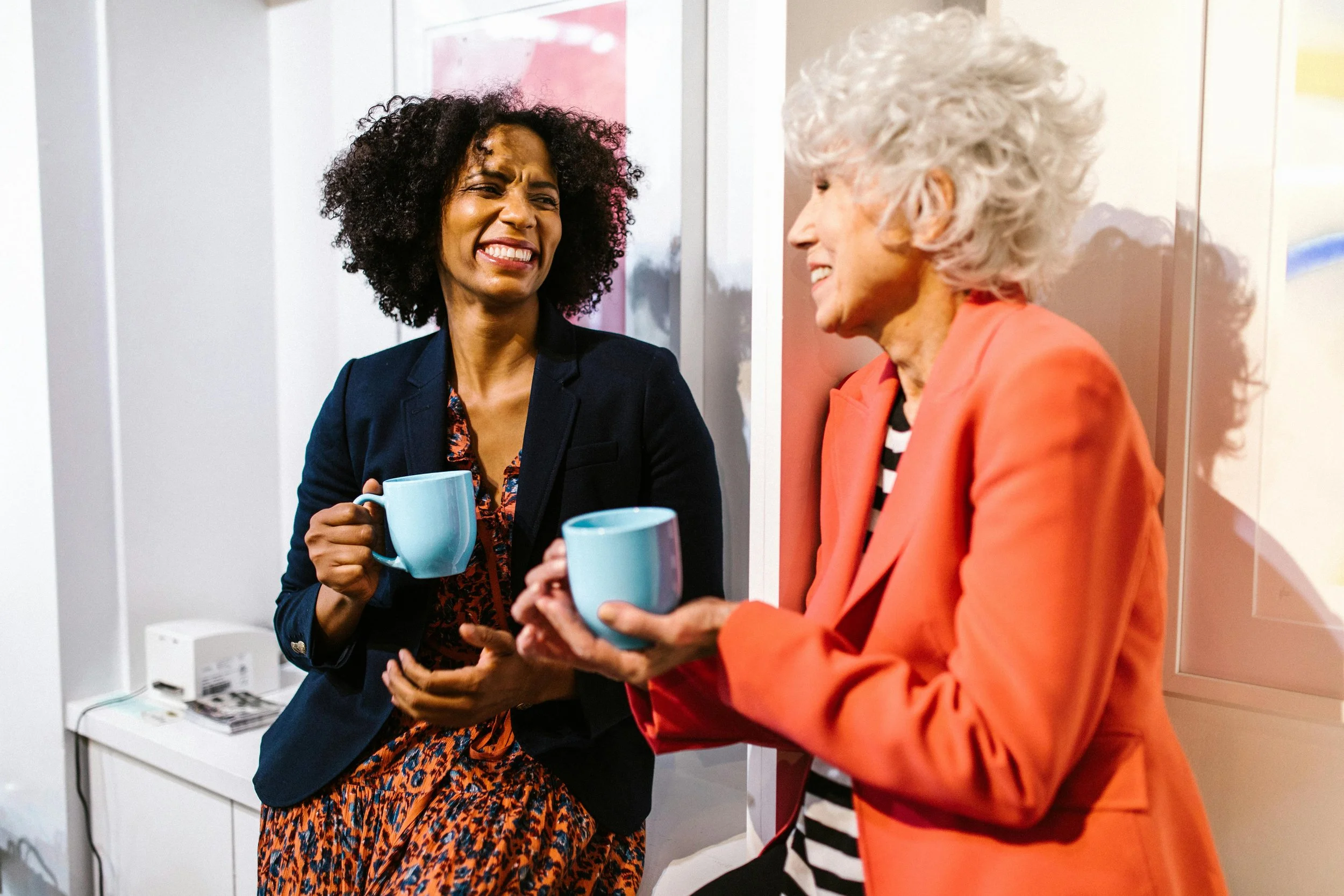 Alt text: Two women are engaged in a lively and warm conversation while holding light blue mugs.