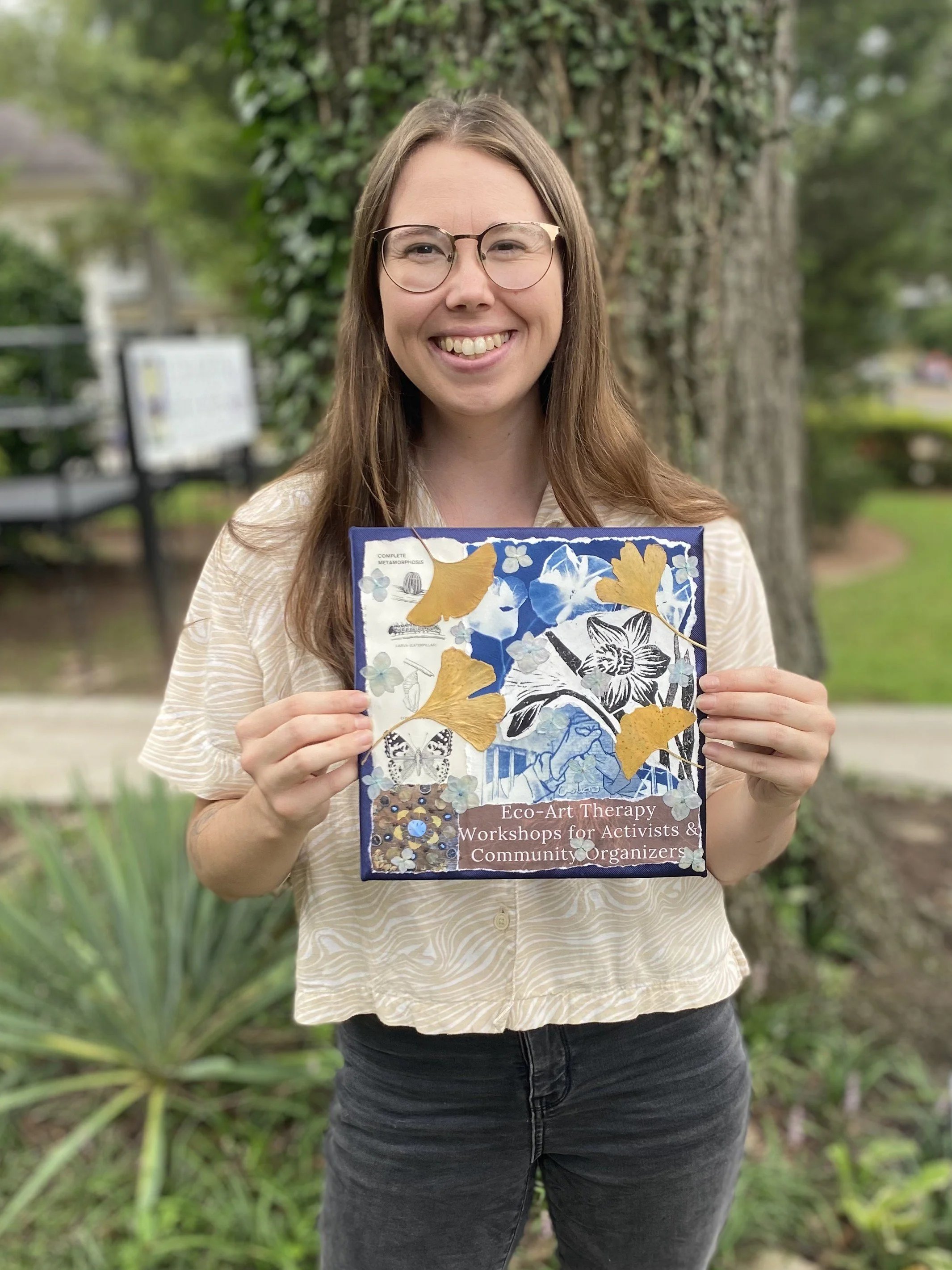 A smiling woman holding a book titled 'Eco-Art Therapy Workshops for Activists & Community Organizers', outdoors in front of a tree with greenery in the background.