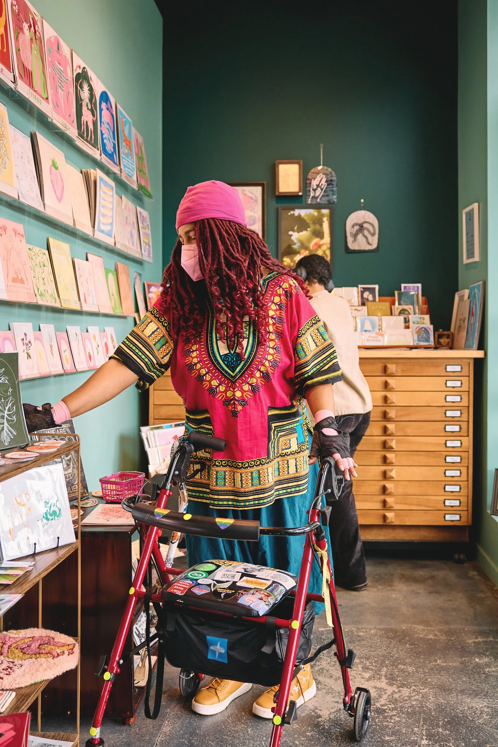 A person with long red dreadlocks, wearing a pink headscarf, pink face mask, colorful dashiki, yellow shoes, and gloves, shopping in an art store with a walker.