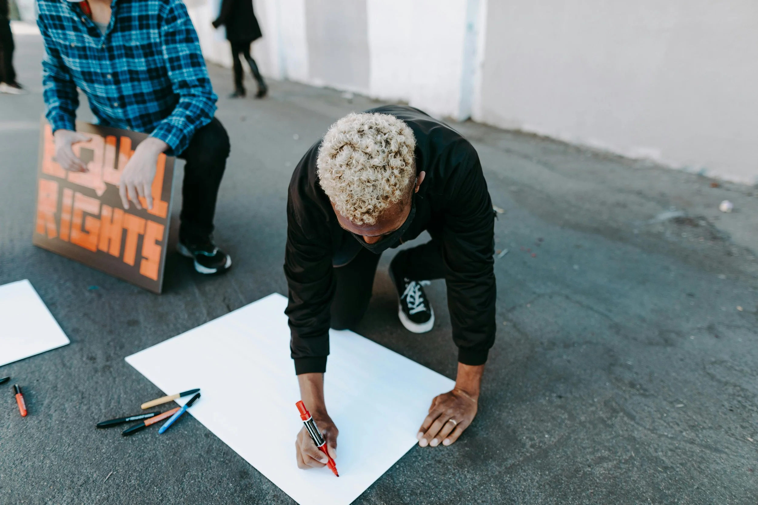 A person with curly blonde hair kneeling on the ground, writing on a large white poster with a red marker. Nearby, there are other markers and a sign that reads 'Black Lives Matter' held by another individual.