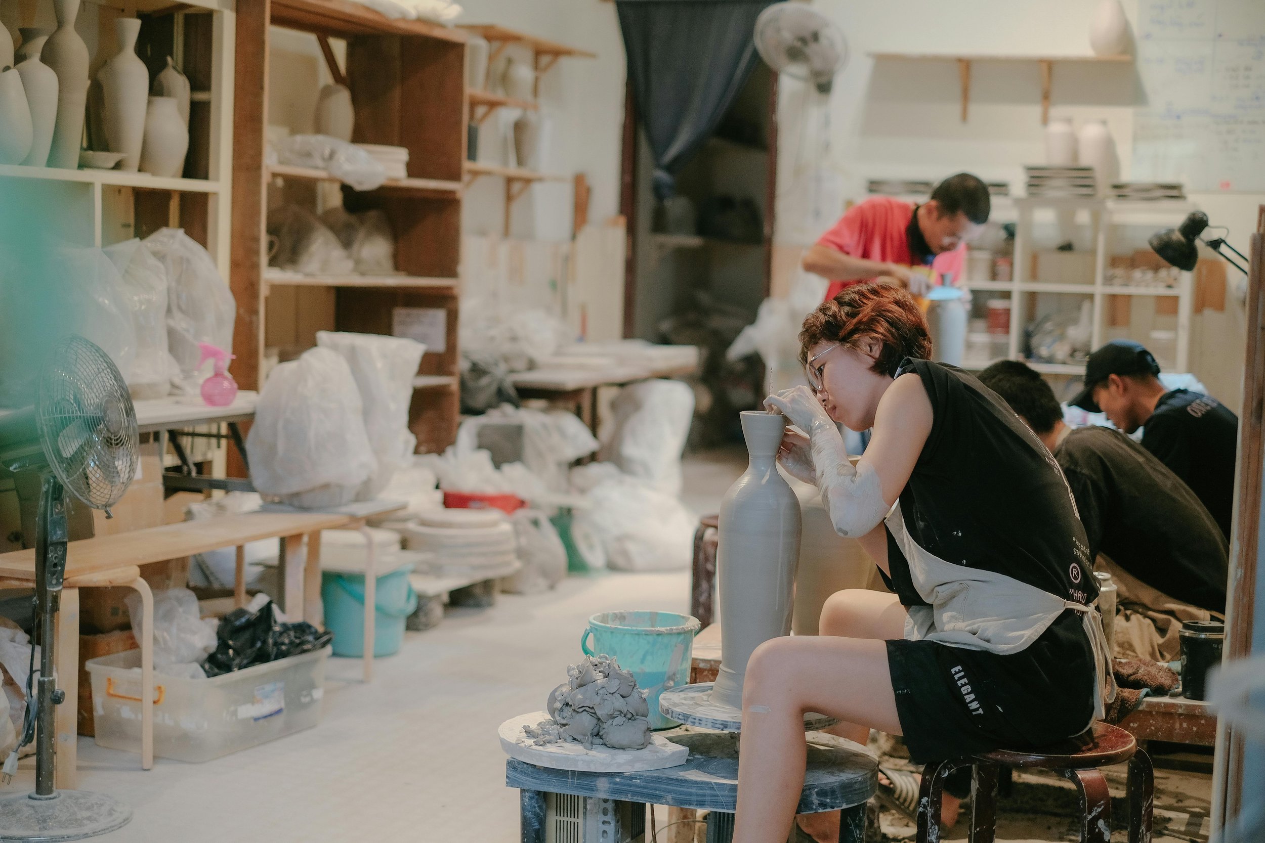 A young woman with glasses sitting on a stool, carefully shaping a ceramic vase on a pottery wheel in a pottery studio surrounded by shelves filled with ceramic pieces and tools.
