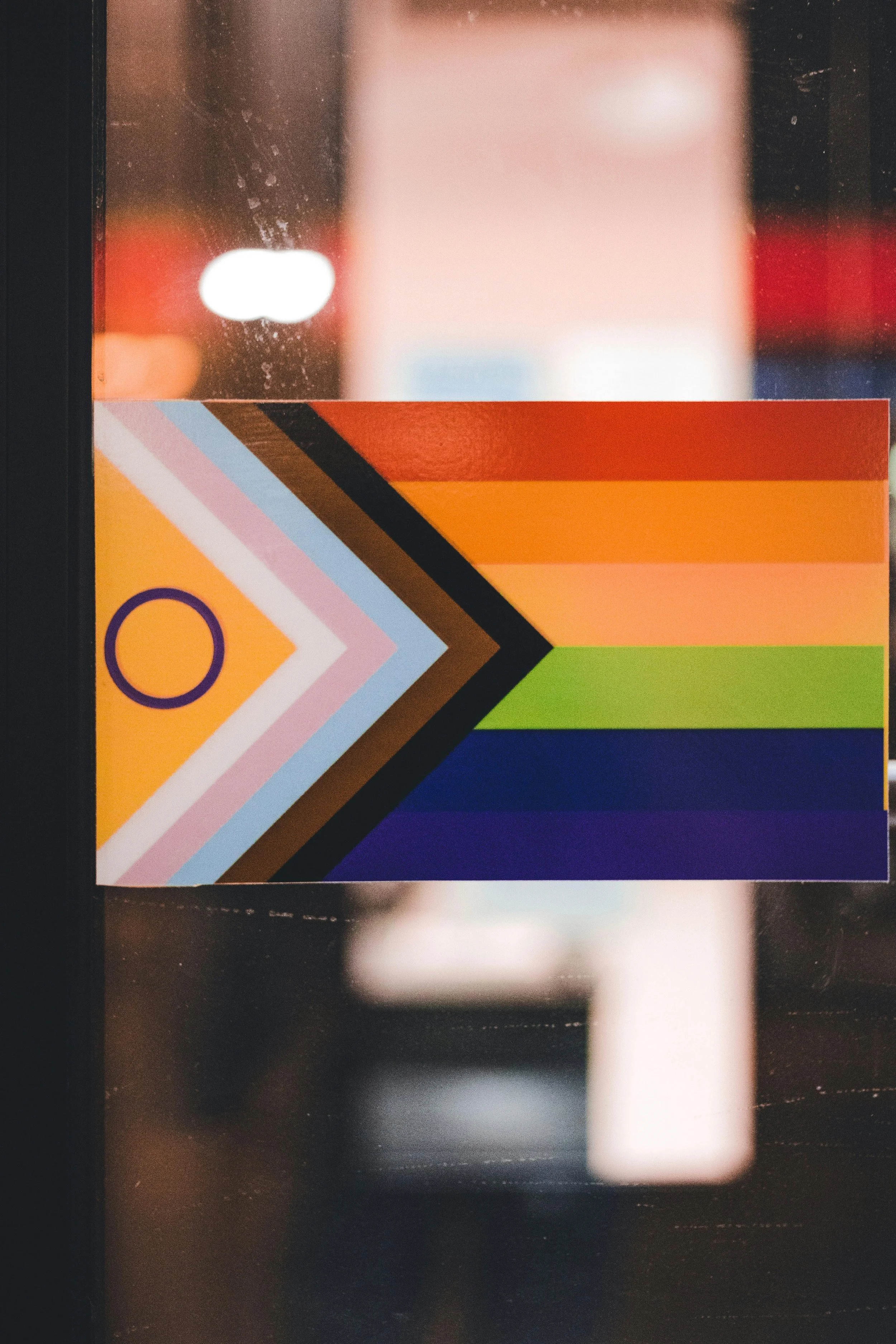 Colorful pride flag with diagonal stripes in orange, peach, yellow, green, blue, and purple, with geometric patterns on the left side.