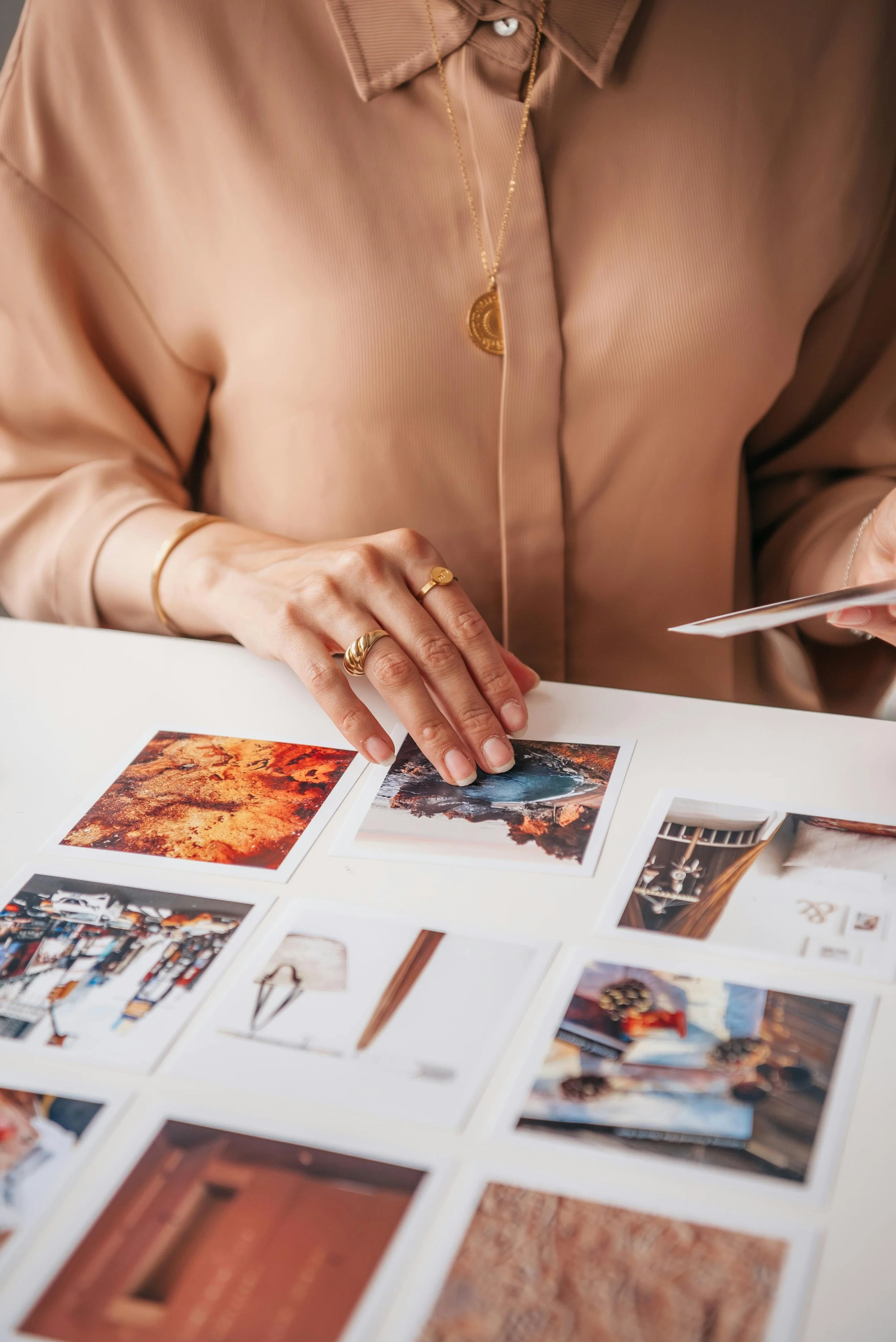 Person wearing a beige silk blouse with gold jewelry, looking at printed photographs laid out on a table.