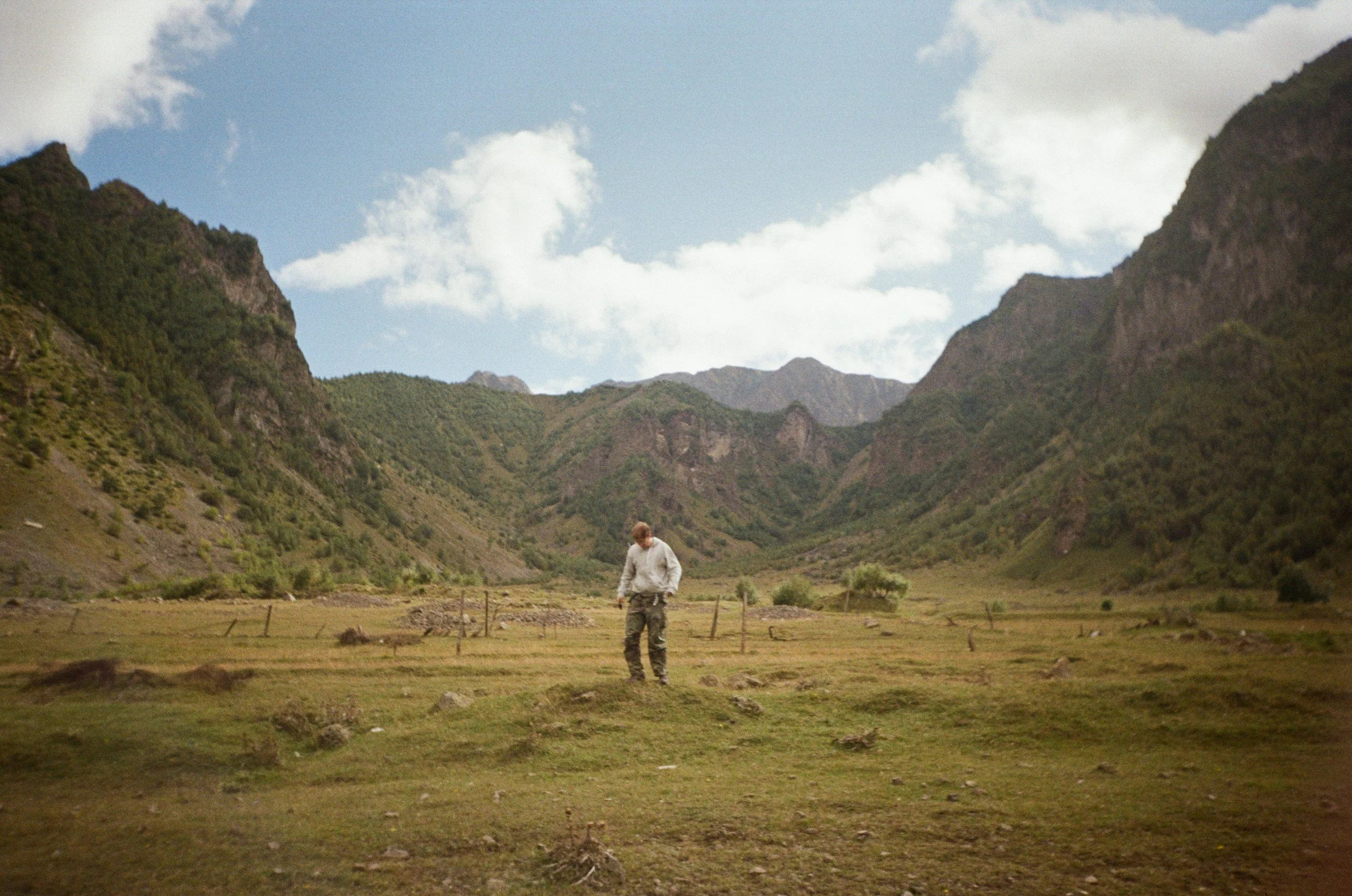 A person walking through a grassy plain surrounded by mountains with clouds in the sky.