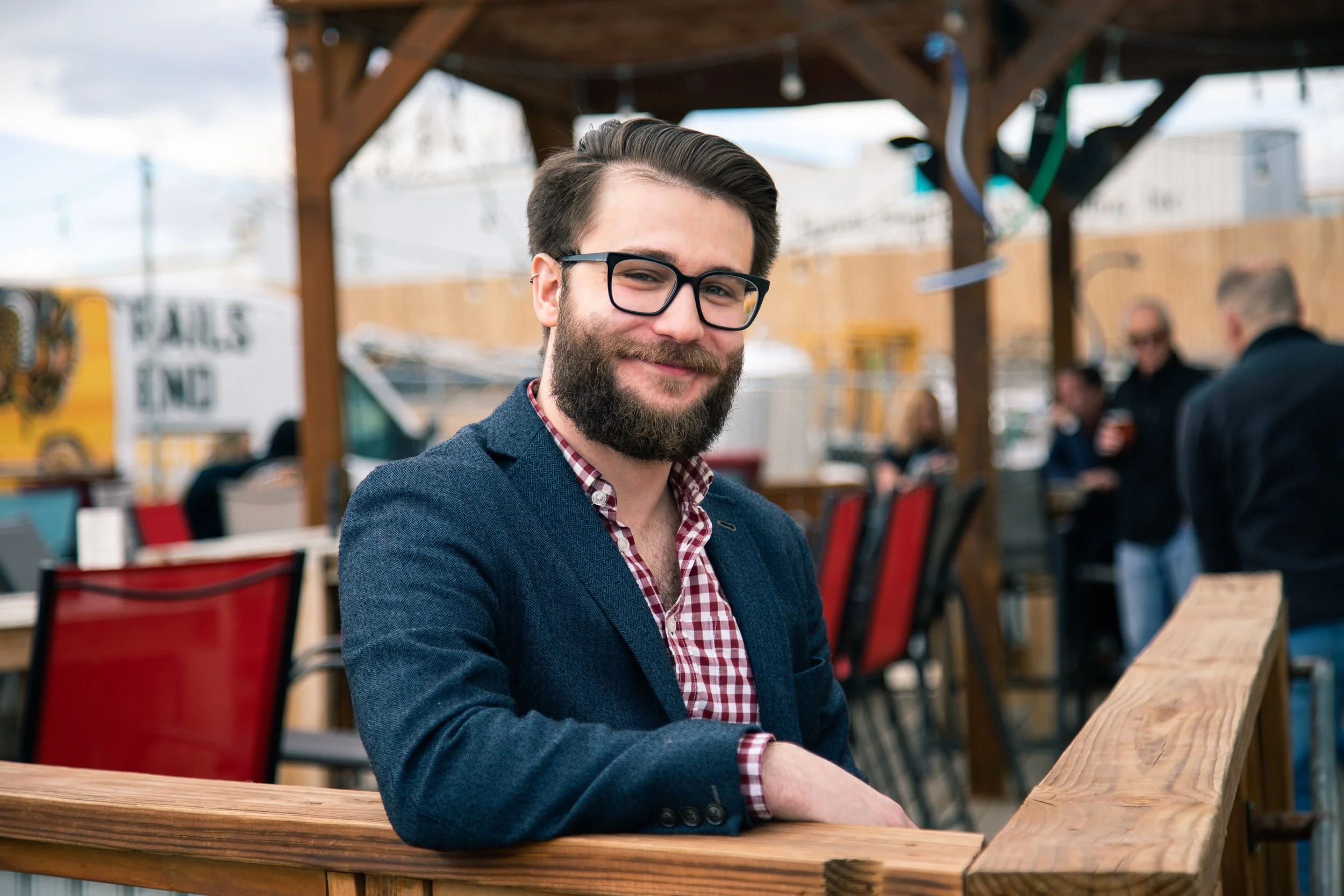 A smiling person with glasses and a beard, wearing a checkered shirt and blazer, sitting at a wooden table outdoors with a blurred background of people and structures.