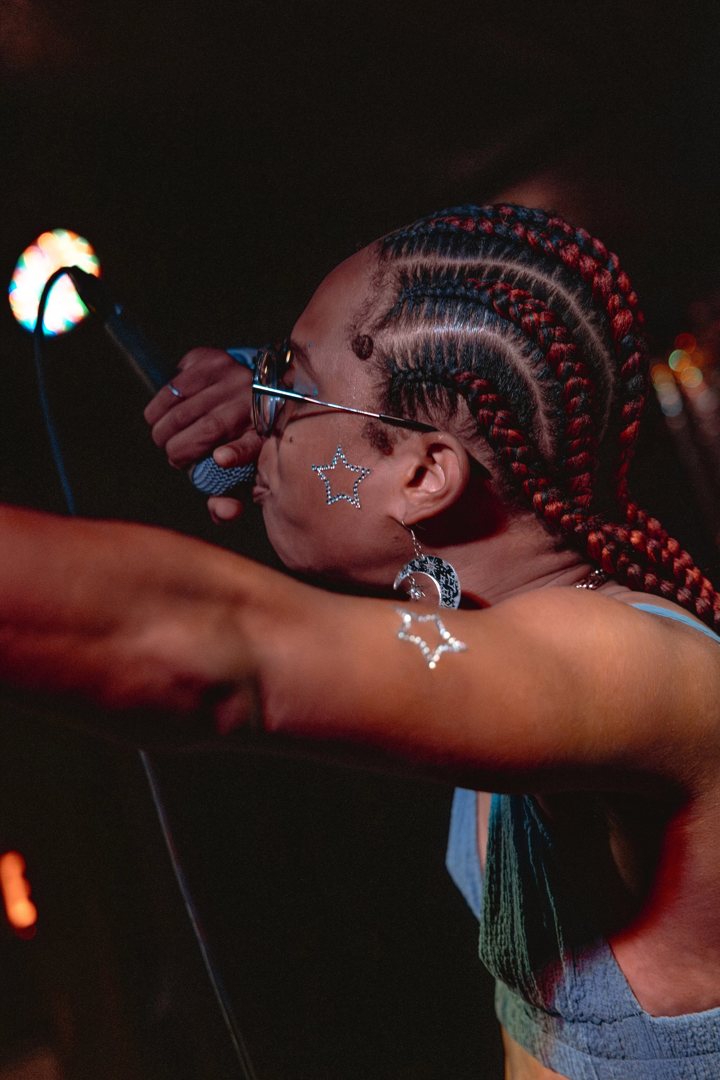 The left side of Erica's face is shown with her left arm, covered in platinum star stickers, extends out to the crowd. She's holding the microphone in her right had as she raps. Her crimson red 6 feed cornrows are braided to the back and she's wearin