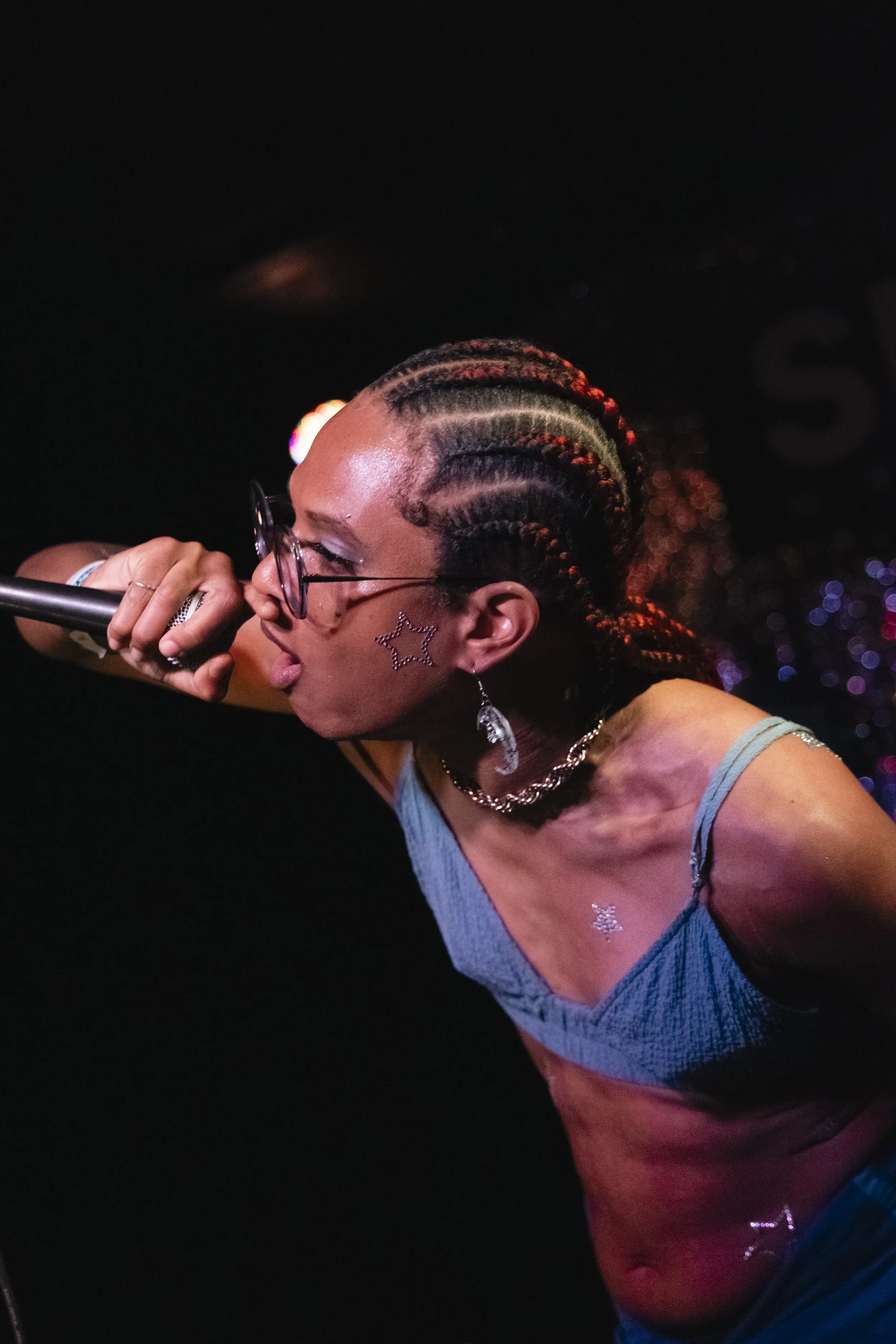 Erica raps with the microphone to her face, wearing thick black rimmed circle shaped glasses, a blue bralette and crimson red braids tied in a bun.