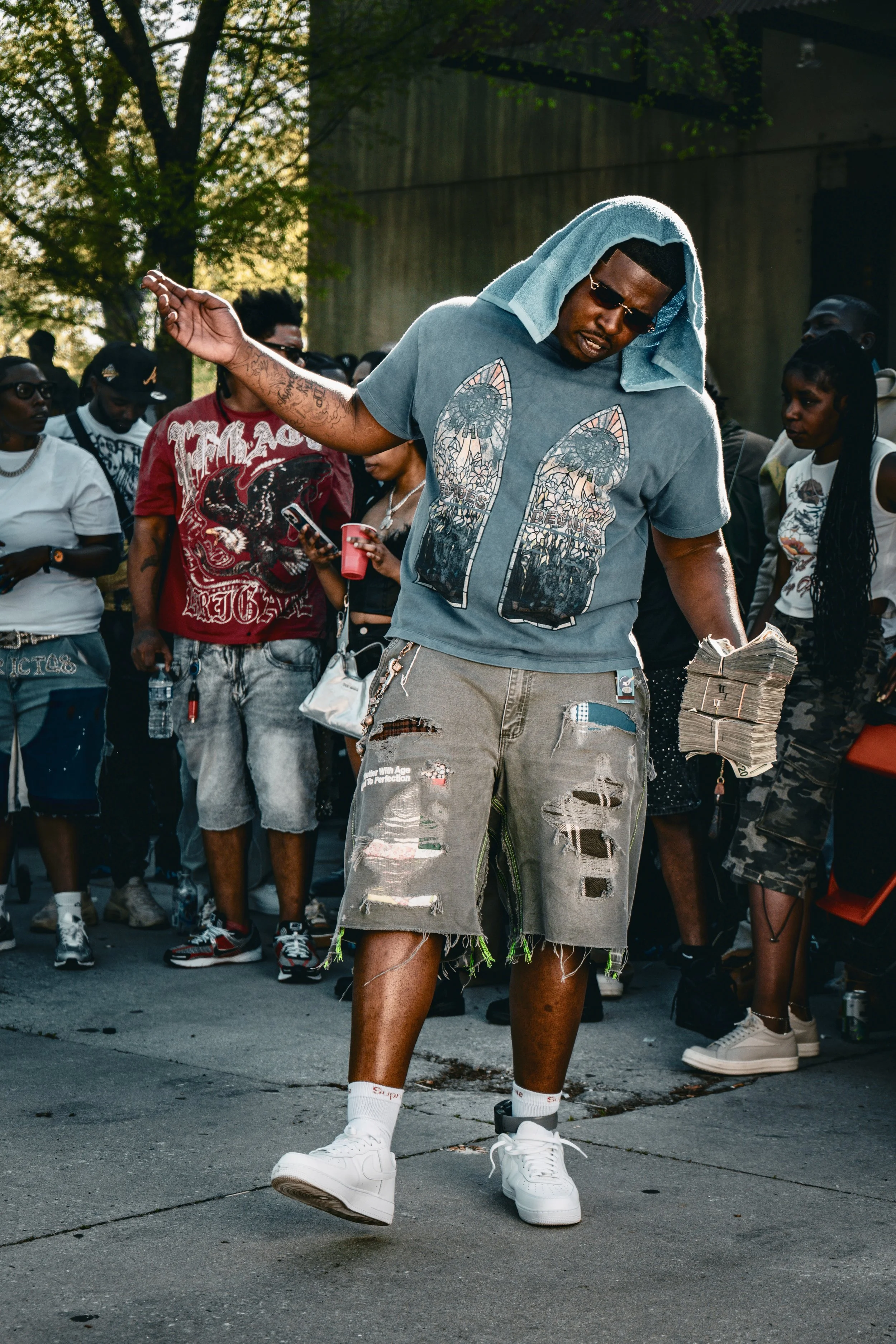 A young man in a gray graphic t-shirt, ripped shorts, and white sneakers participates in an outdoor event, with a towel over his head and a stack of money in his hand. Several people are gathered around in casual attire, some taking photos or observi
