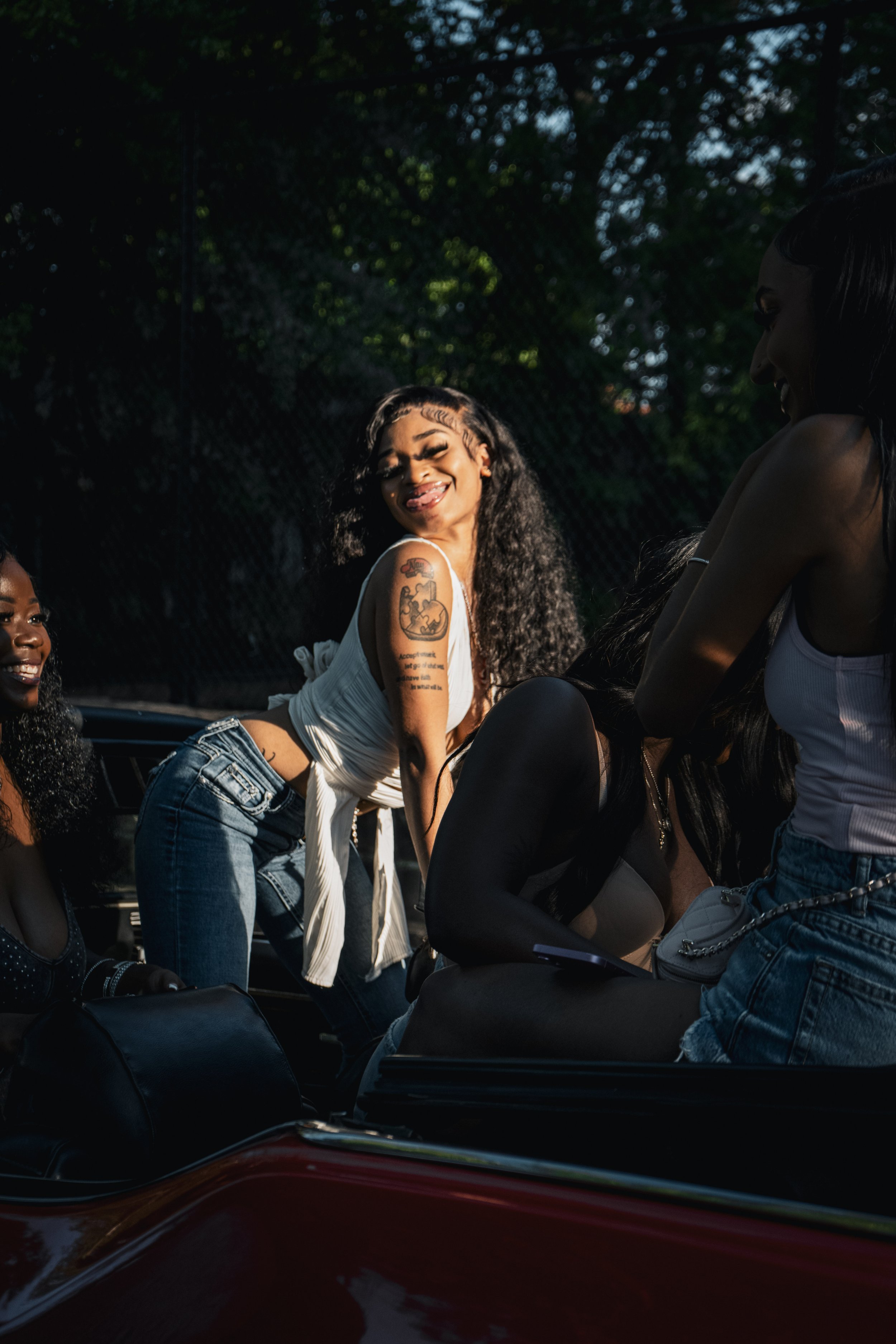 Group of young women sitting and standing outdoors, smiling, with a dark green leafy background, dressed casually.