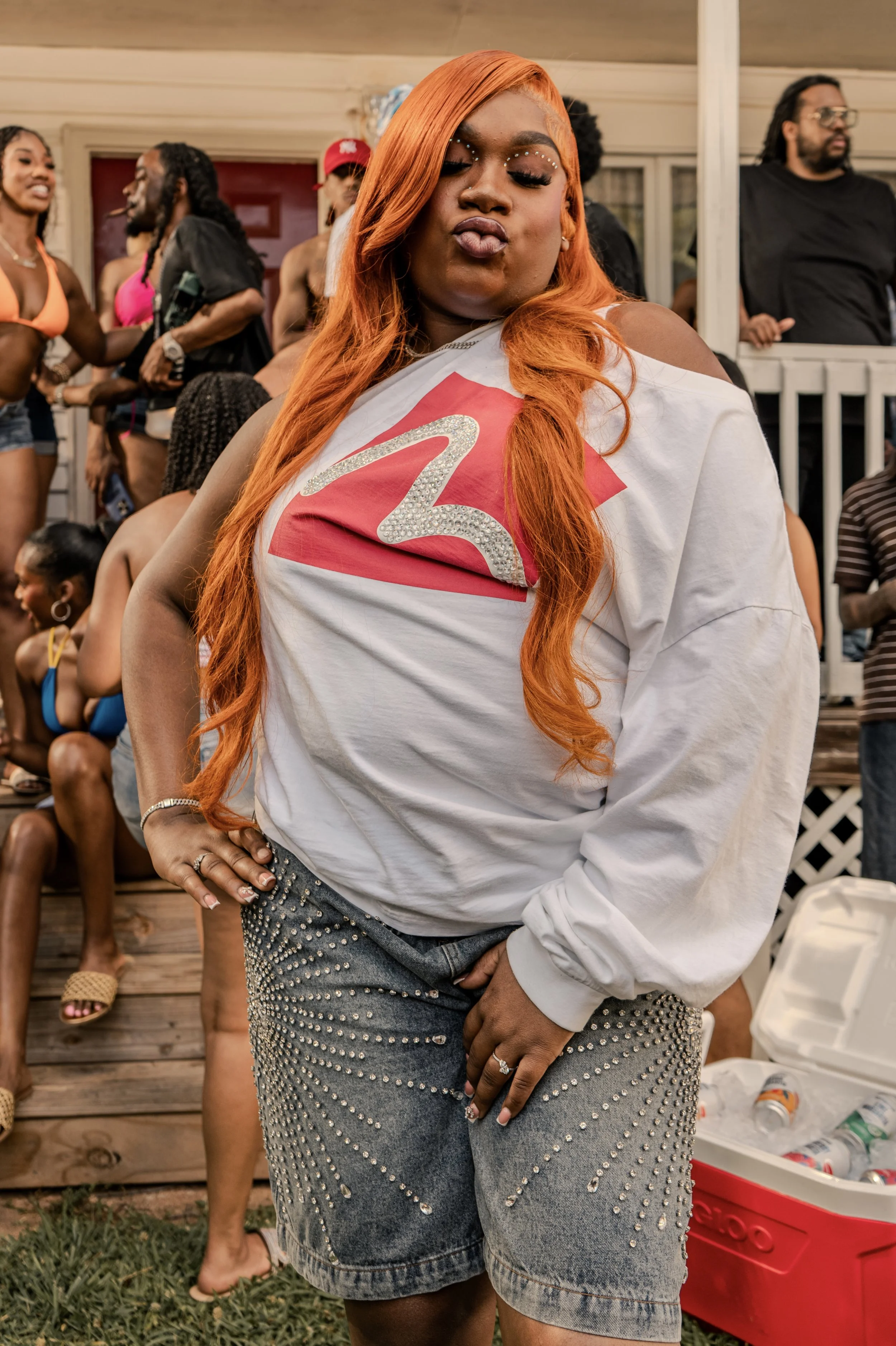 A woman with long orange hair making a duck face, wearing a white t-shirt with a large glittery number 2 on it and distressed jeans adorned with silver studs at a social gathering.