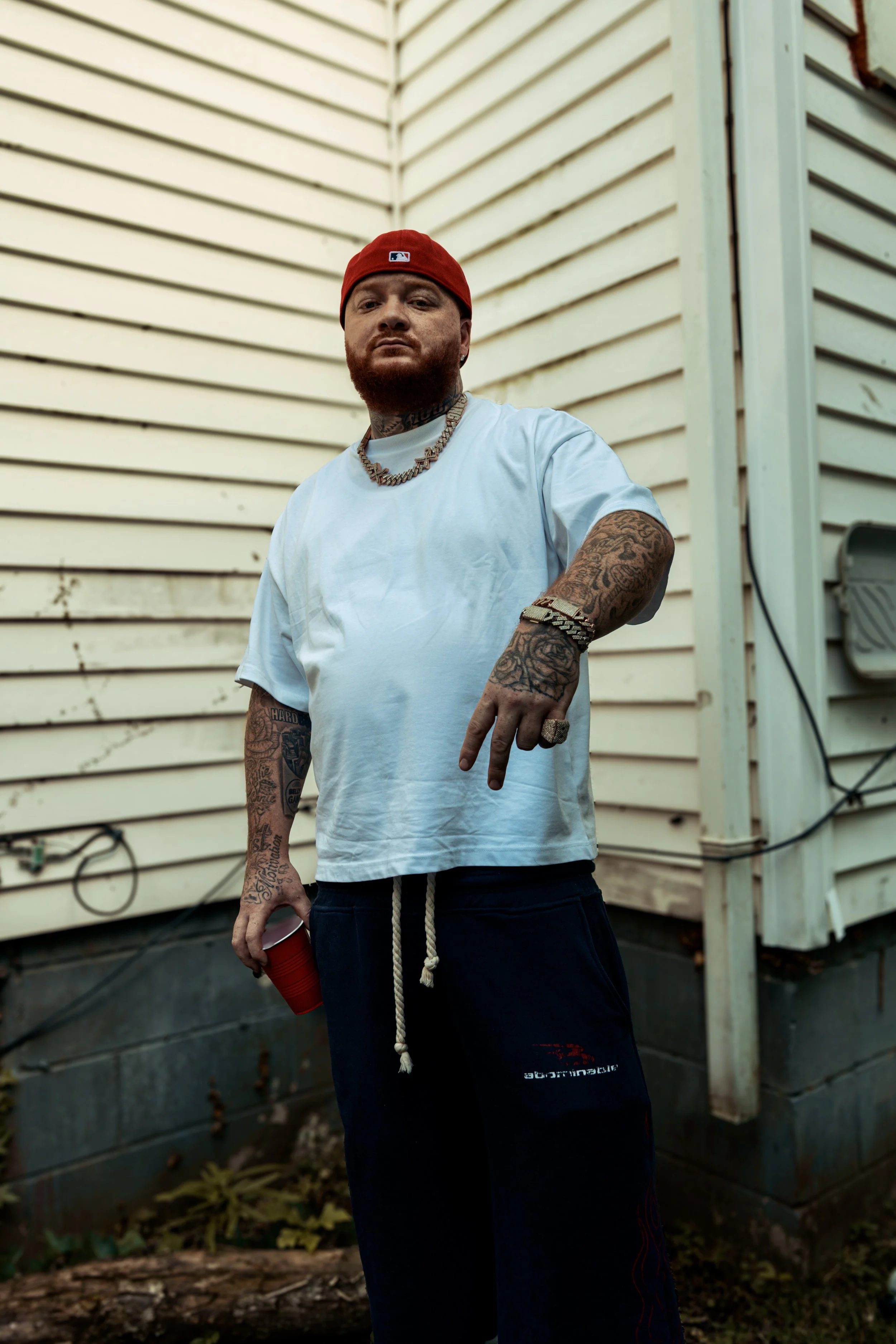 A man with tattoos wearing a red cap, white t-shirt, and black shorts holding a red cup outside a house with beige siding and visible electrical wiring.