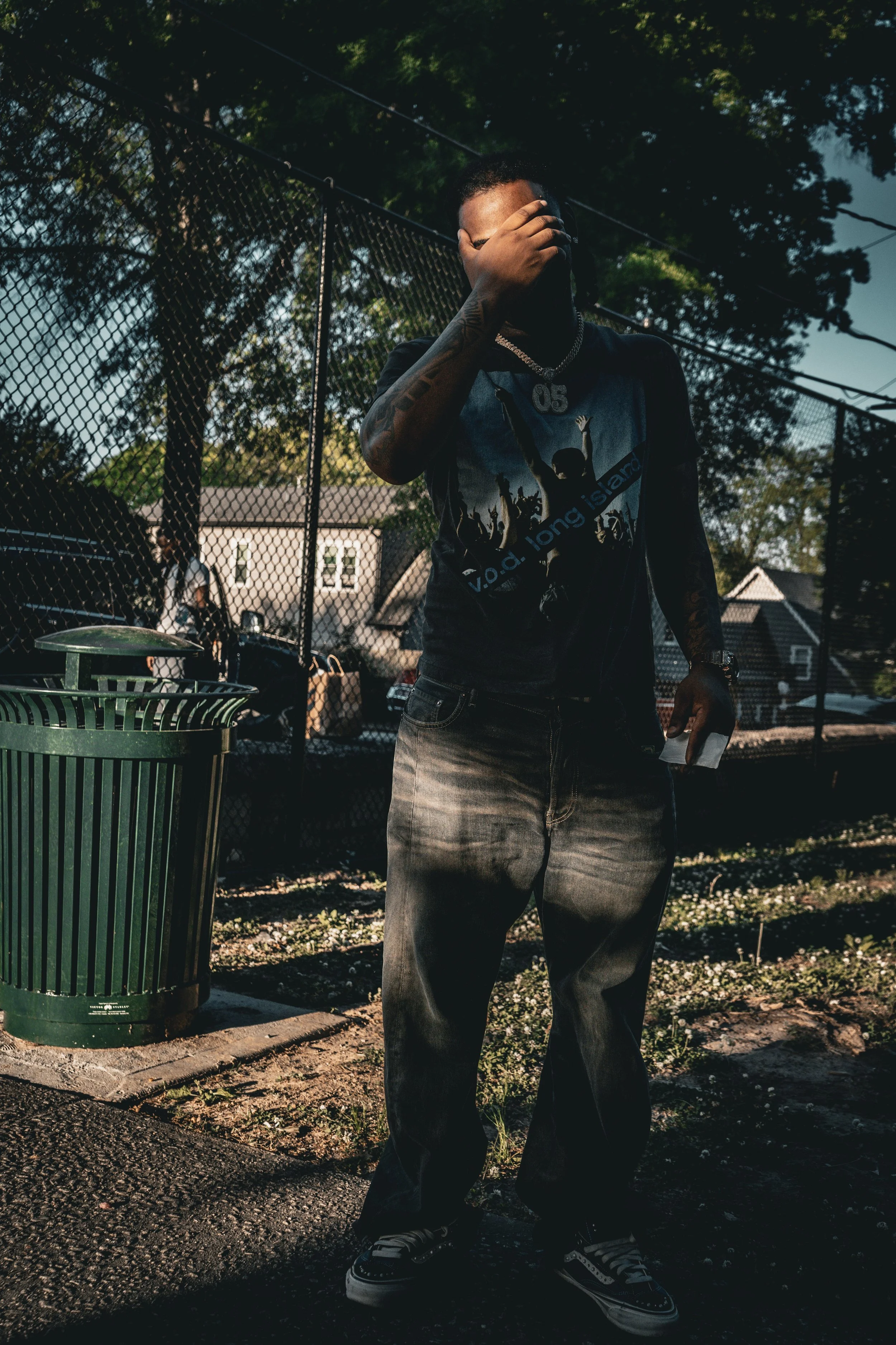 A man standing outdoors near a chain-link fence, covering his face with his hand. He is wearing a graphic t-shirt with a design featuring people, jeans, sneakers, a chain necklace, and a wristwatch. A person can be seen in the background behind the f