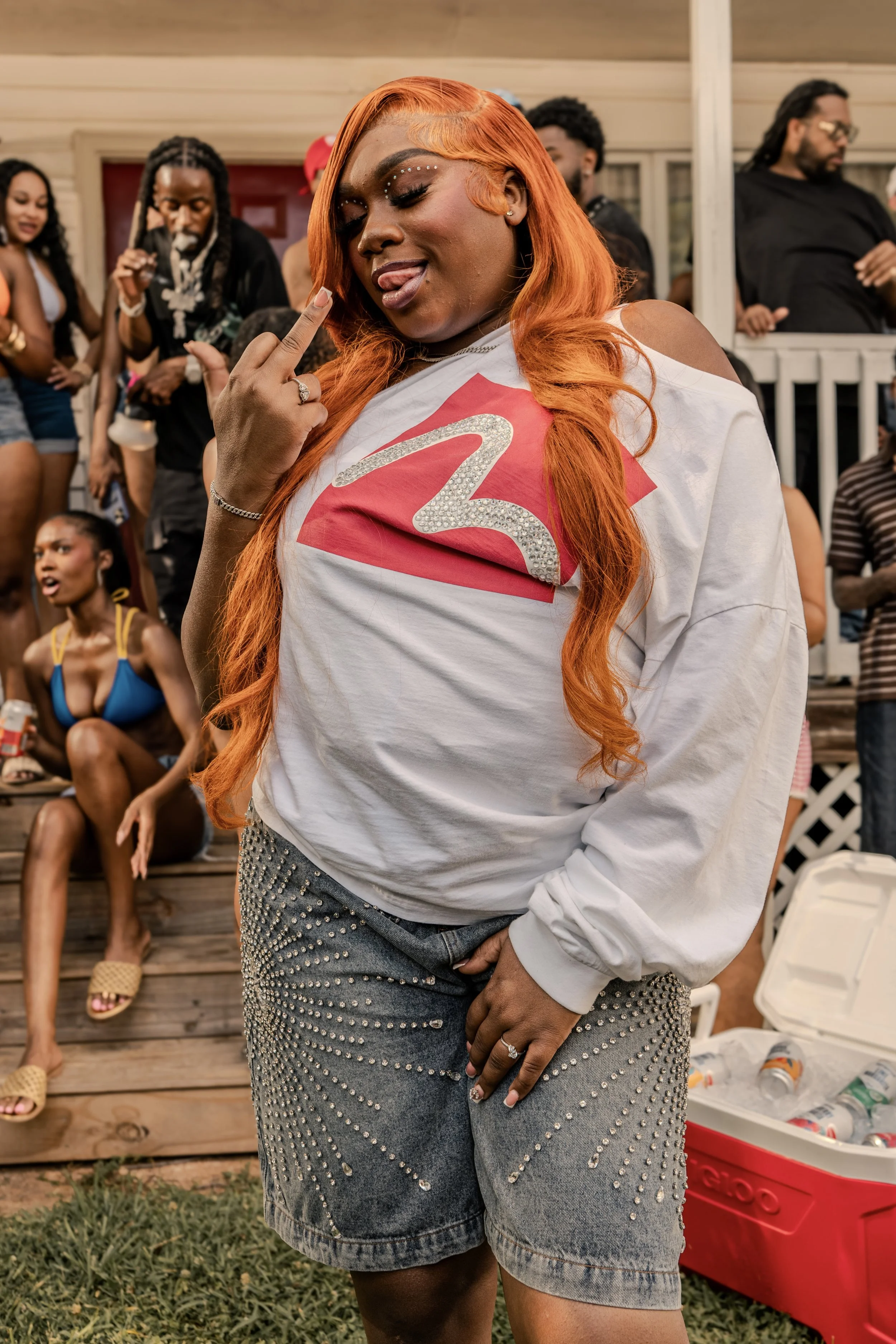 Woman with orange hair and rhinestone makeup sticking out her tongue at a social gathering, standing in front of a group of diverse people on a porch.