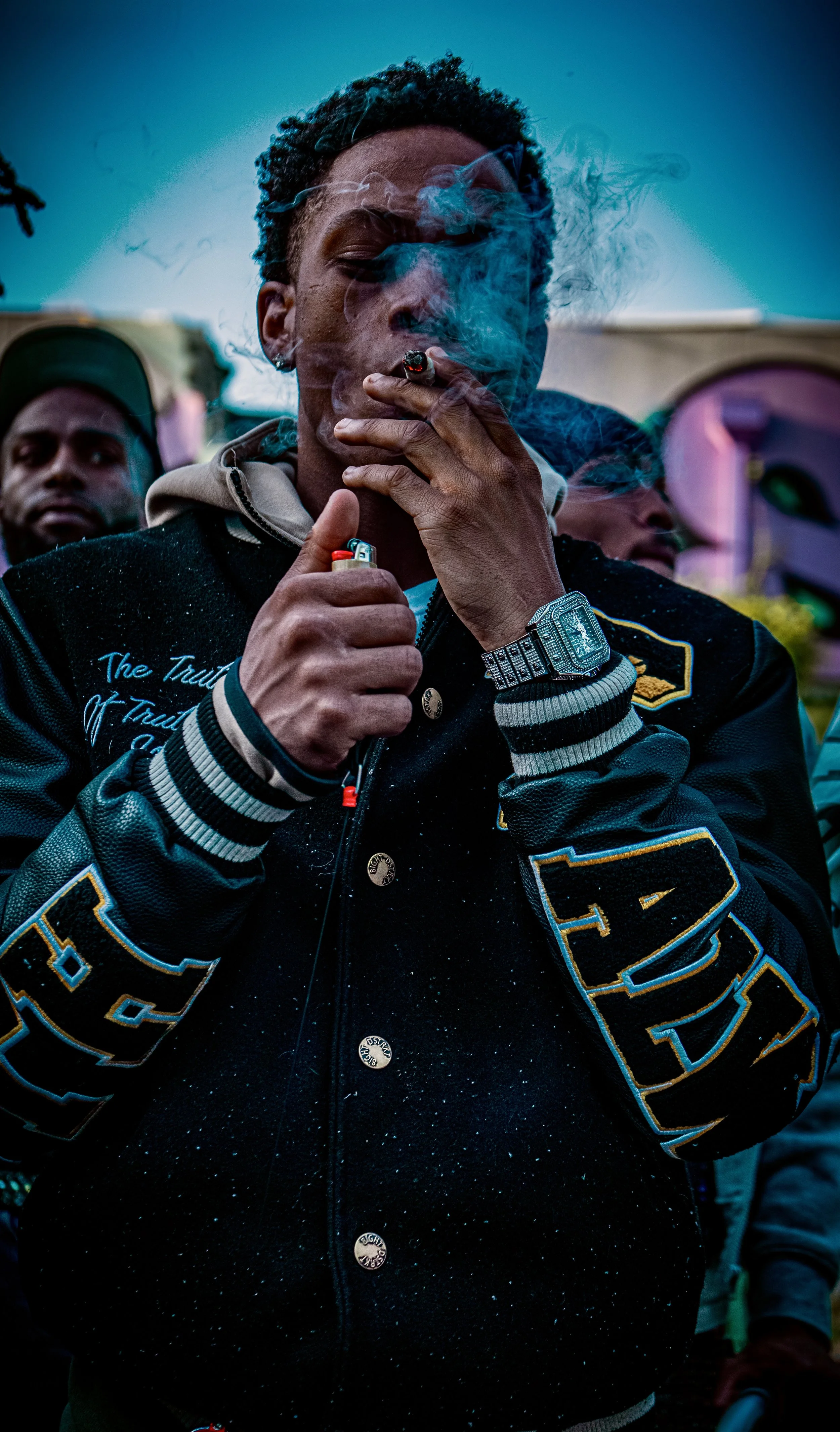 A young man with curly hair wearing a black varsity jacket with blue and yellow embroidery, a silver watch, and earring, smoking a cigarette, surrounded by others.